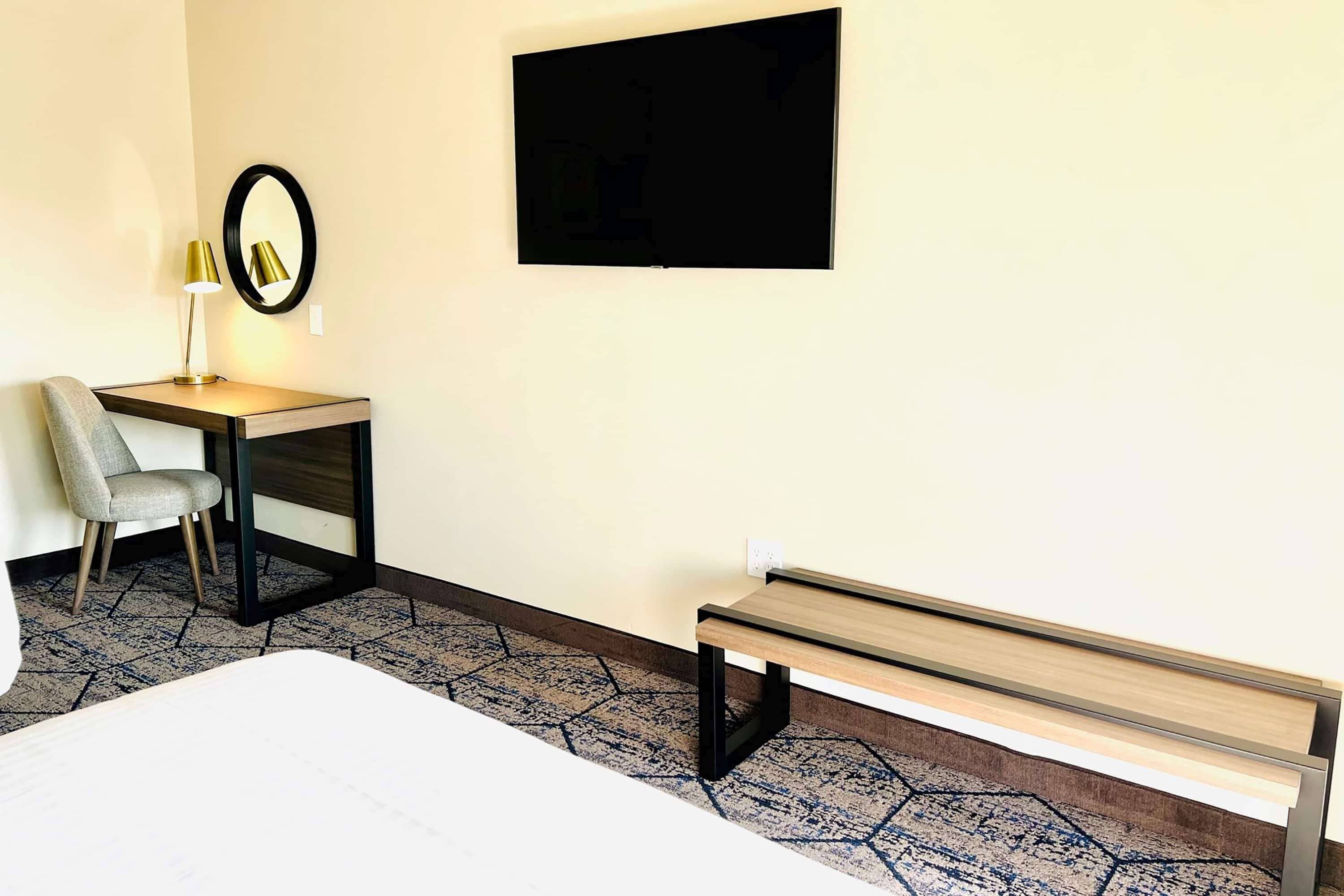 Habitación de hotel con cama, escritorio y silla, banca, televisor montado en la pared y alfombra estampada.