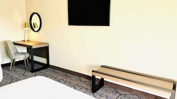 Habitación de hotel con cama, escritorio y silla, banca, televisor montado en la pared y alfombra estampada.