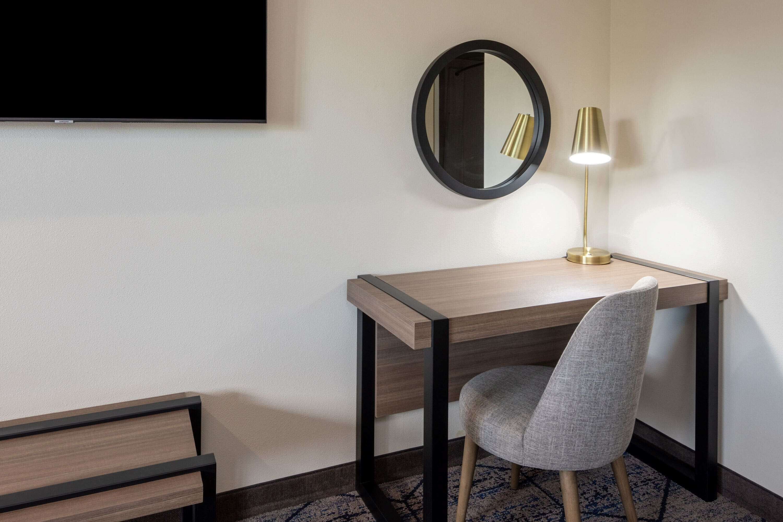 Habitació d'hotel amb escriptori de fusta, cadira tapissada de color gris, mirall rodó i làmpada de peu.