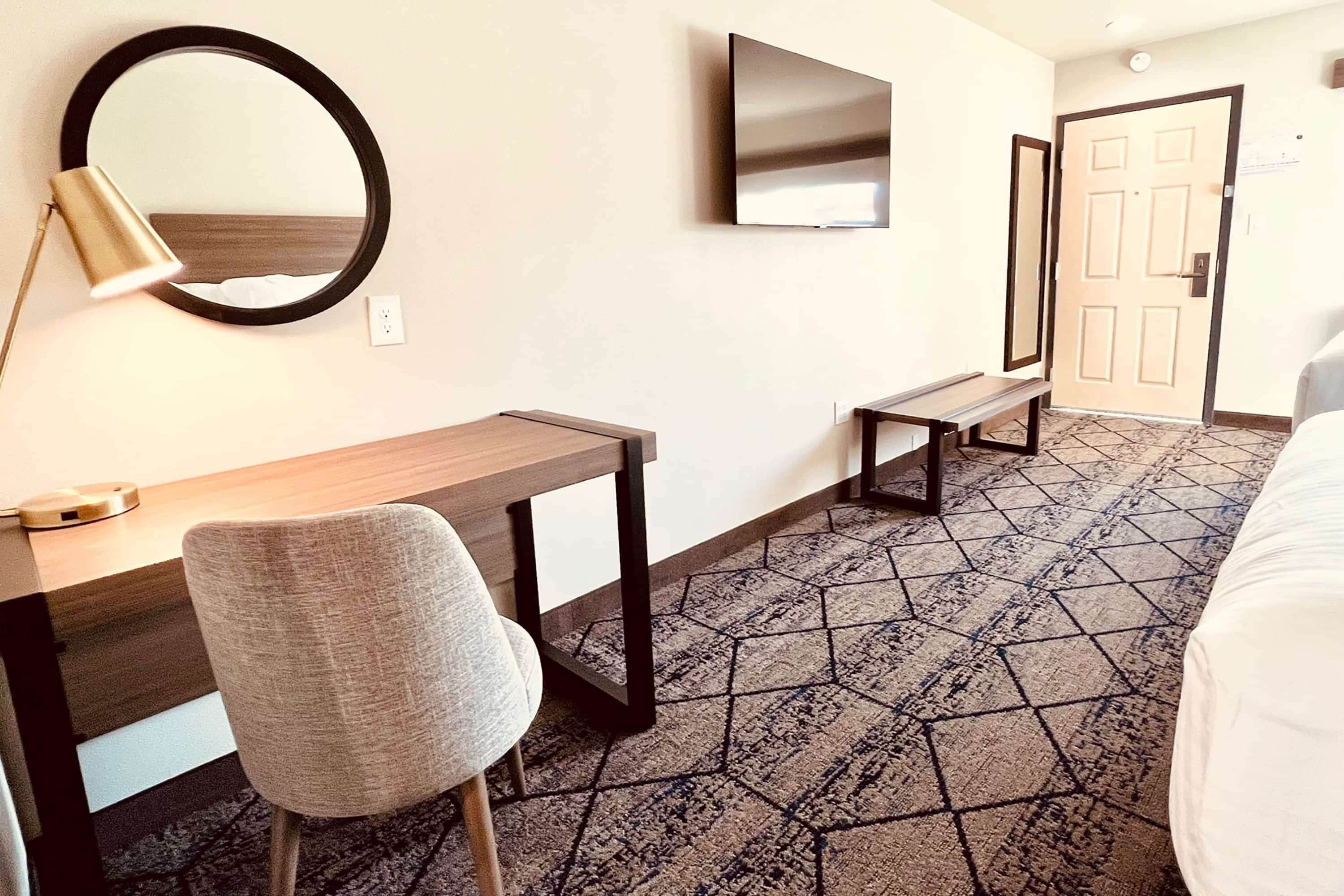 A hotel room with a bed, a desk, a chair, a round mirror, a television, and a door.