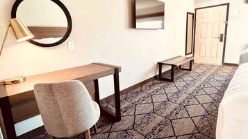 A hotel room with a bed, a desk, a chair, a round mirror, a television, and a door.