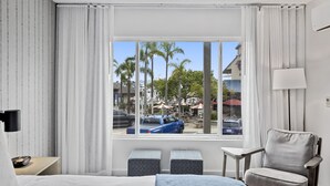 A hotel room with a large bed, a chair, a small table, and a window with a view of palm trees and parked cars outside. - Sea Harbor Hotel (San Diego)