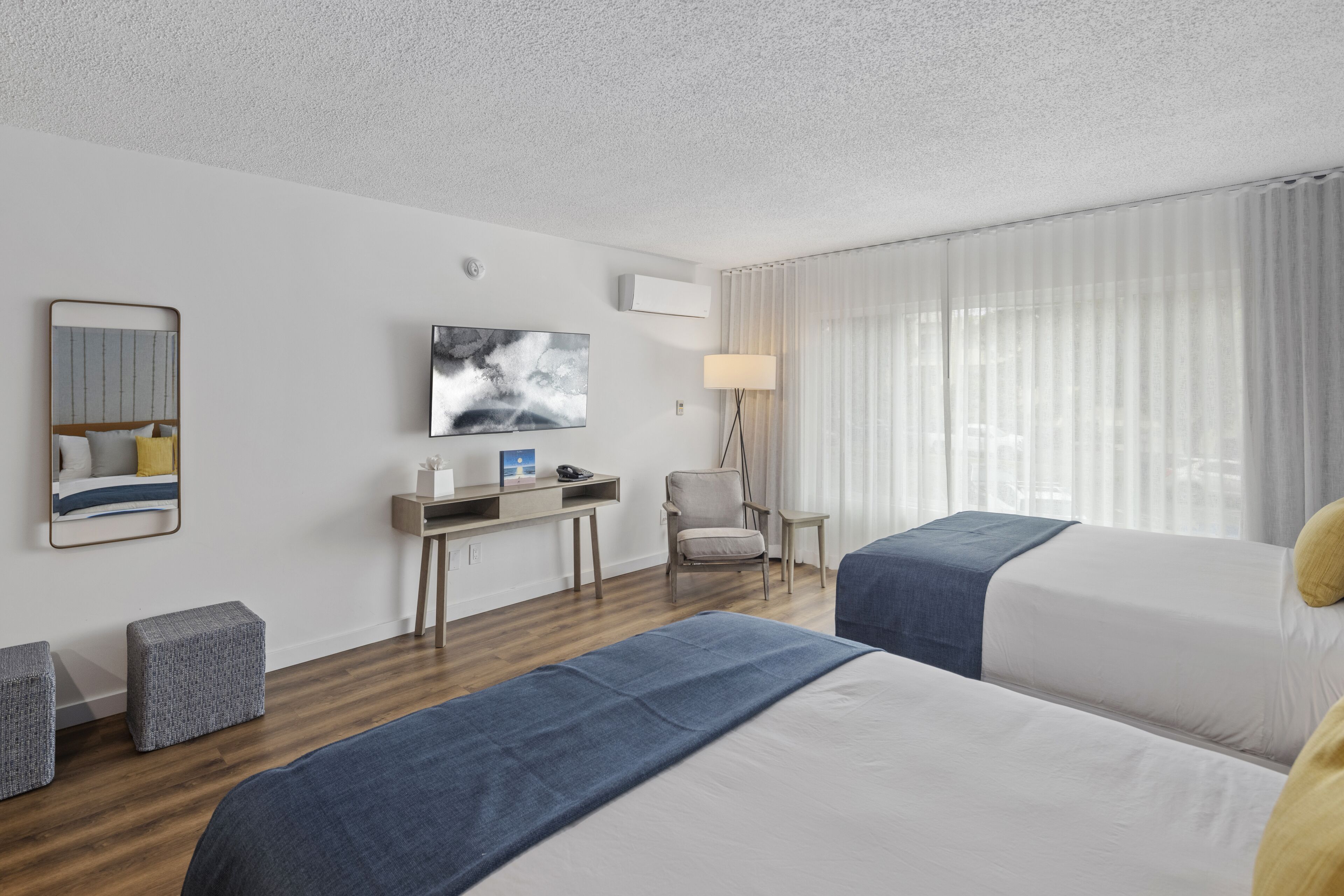 Habitación de hotel con dos camas, un escritorio, una silla, una lámpara y un ventanal grande con cortinas.
