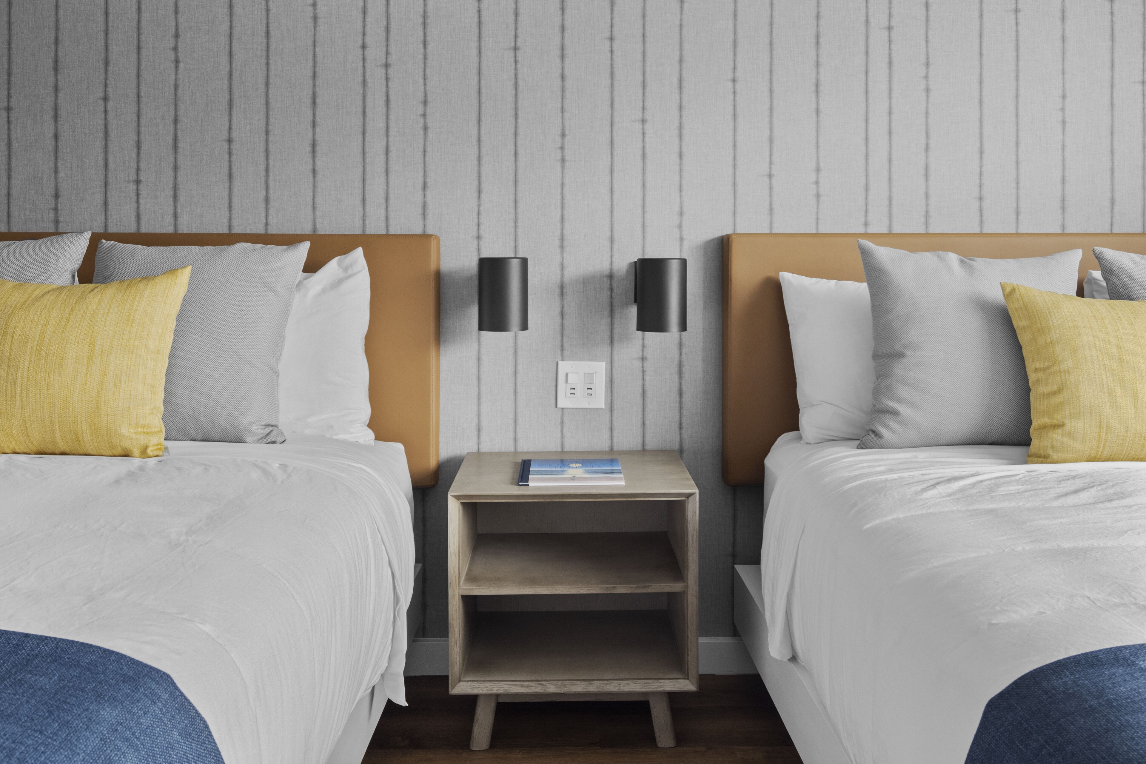 Quarto de hotel com duas camas, uma mesa de cabeceira com um livro e um candeeiro fixo na parede.