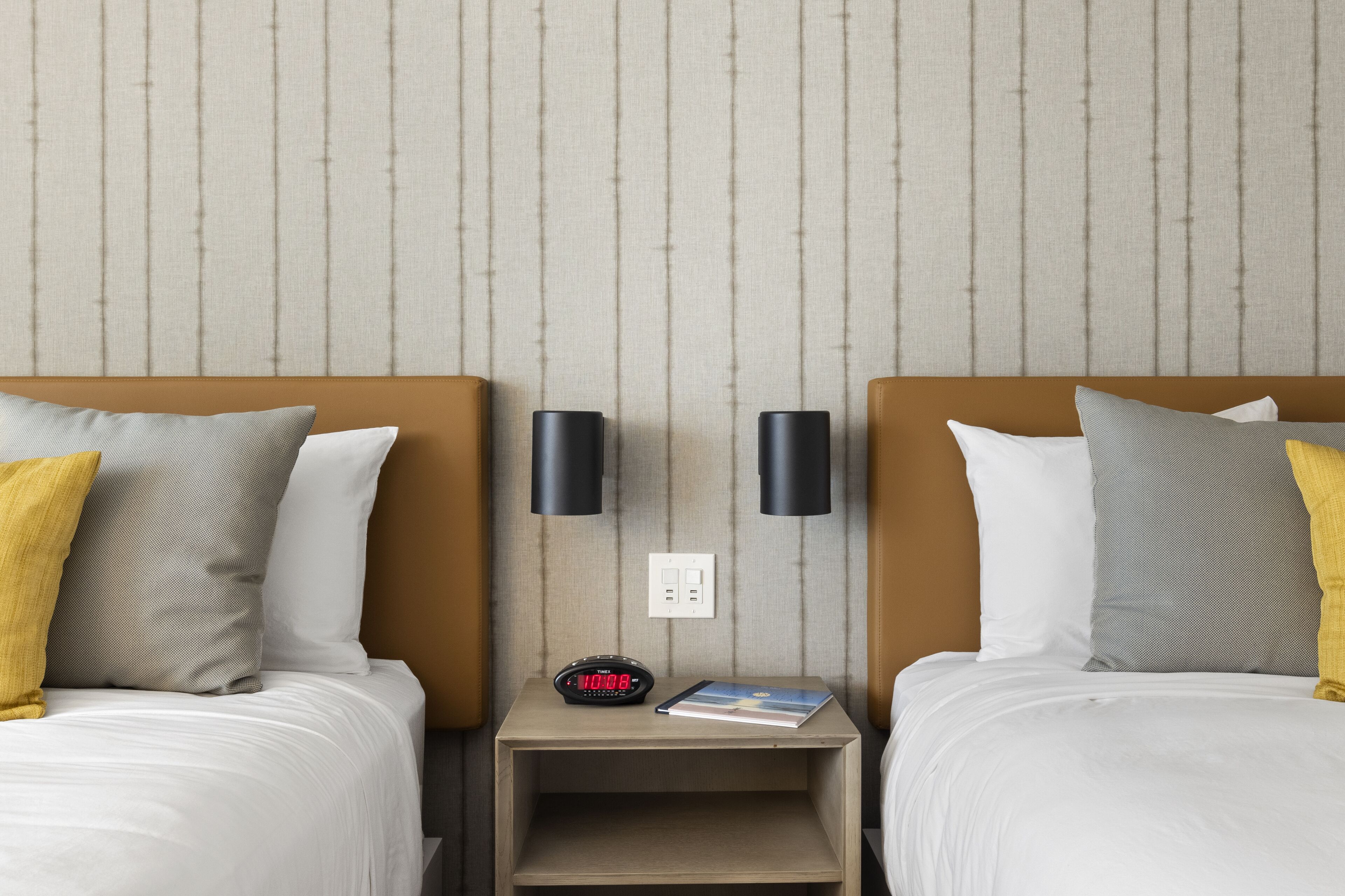Ein Hotelzimmer mit zwei Betten, jedes mit einem Kopfkissen, einem Nachttisch mit einer Uhr und einem Buch sowie zwei Wandleuchten.