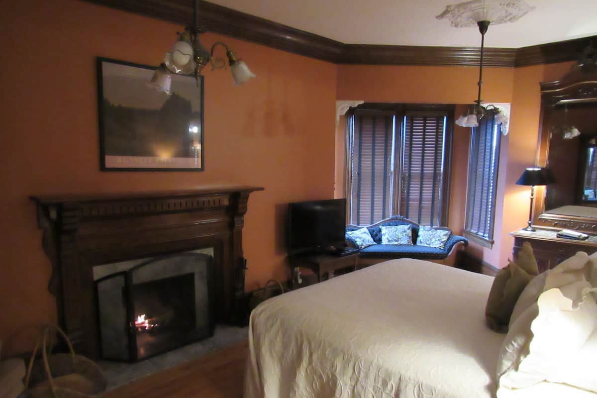A bedroom with a fireplace, a bed, a television, and a window with shutters.