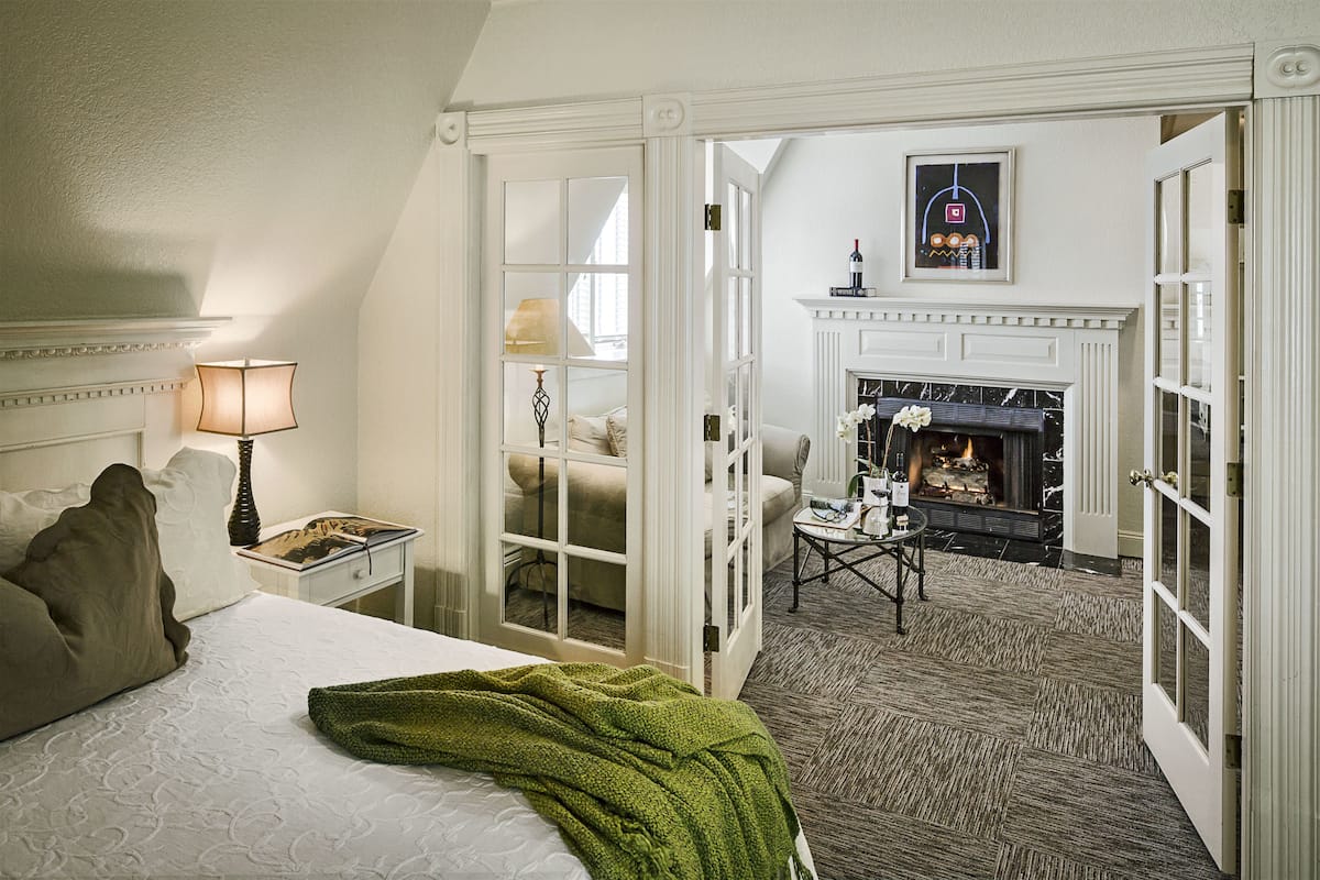 A hotel room with a bed, a nightstand, a lamp, a green throw blanket, a chair, a small table, and a fireplace in the background.