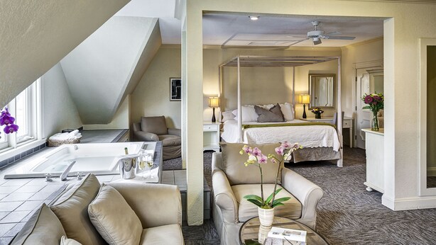 A hotel room with a bed, a sitting area with a sofa and armchairs, a small table with a vase of flowers, and a bathroom with a bathtub and shower. - Carter House Inns (Eureka)