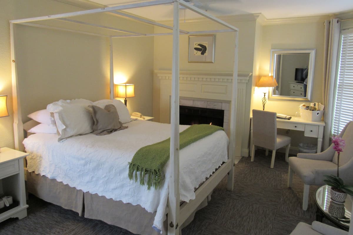 A four-poster bed with a green throw blanket, a desk with a chair, a mirror, and a small potted plant.