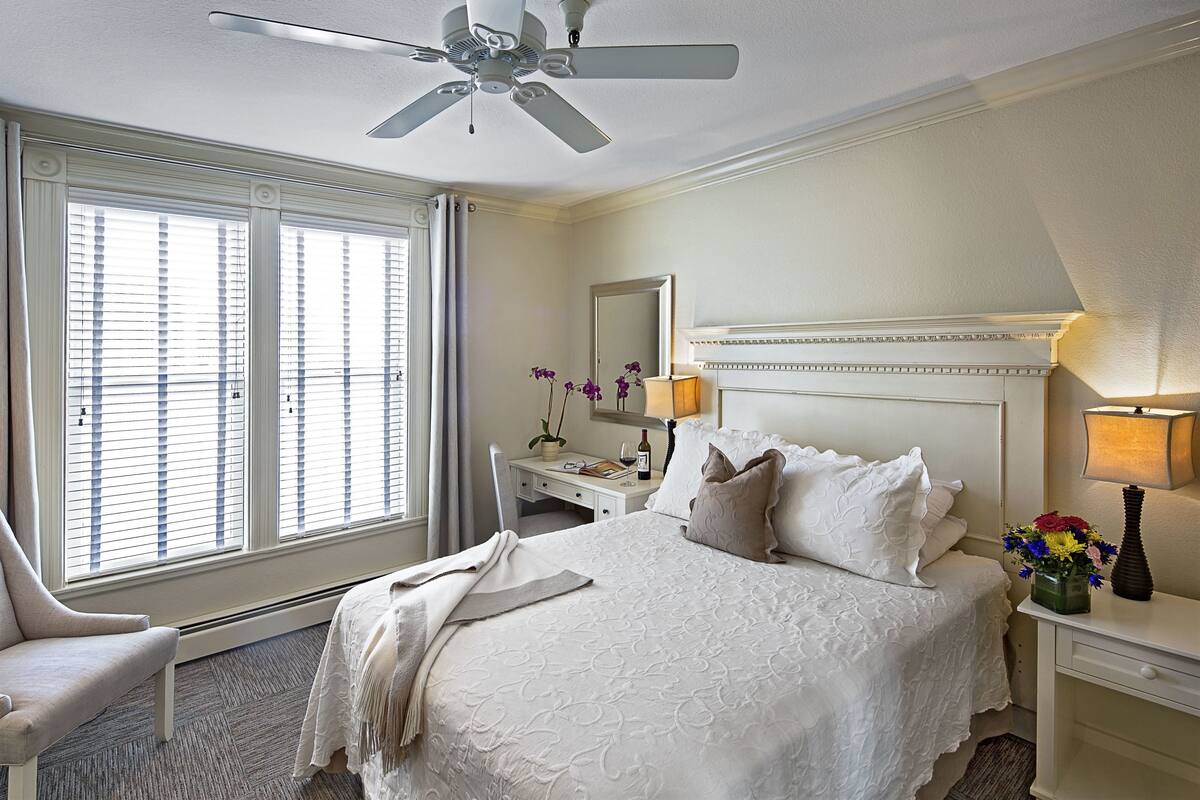 A hotel room with a large bed, a desk, a chair, a ceiling fan, a window with blinds, and a nightstand with a lamp and flowers.