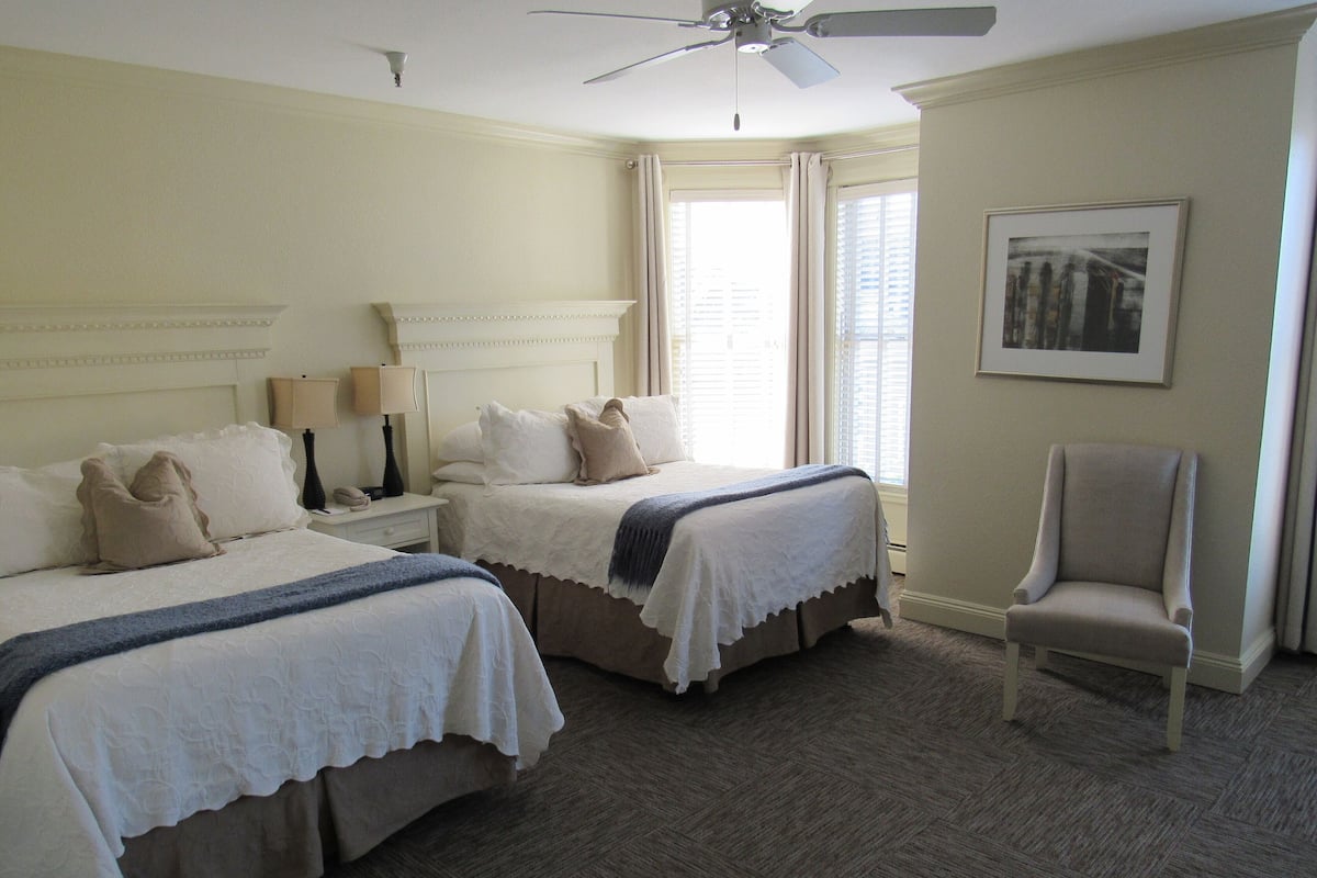A hotel room with two beds, a chair, a ceiling fan, and a window with curtains.
