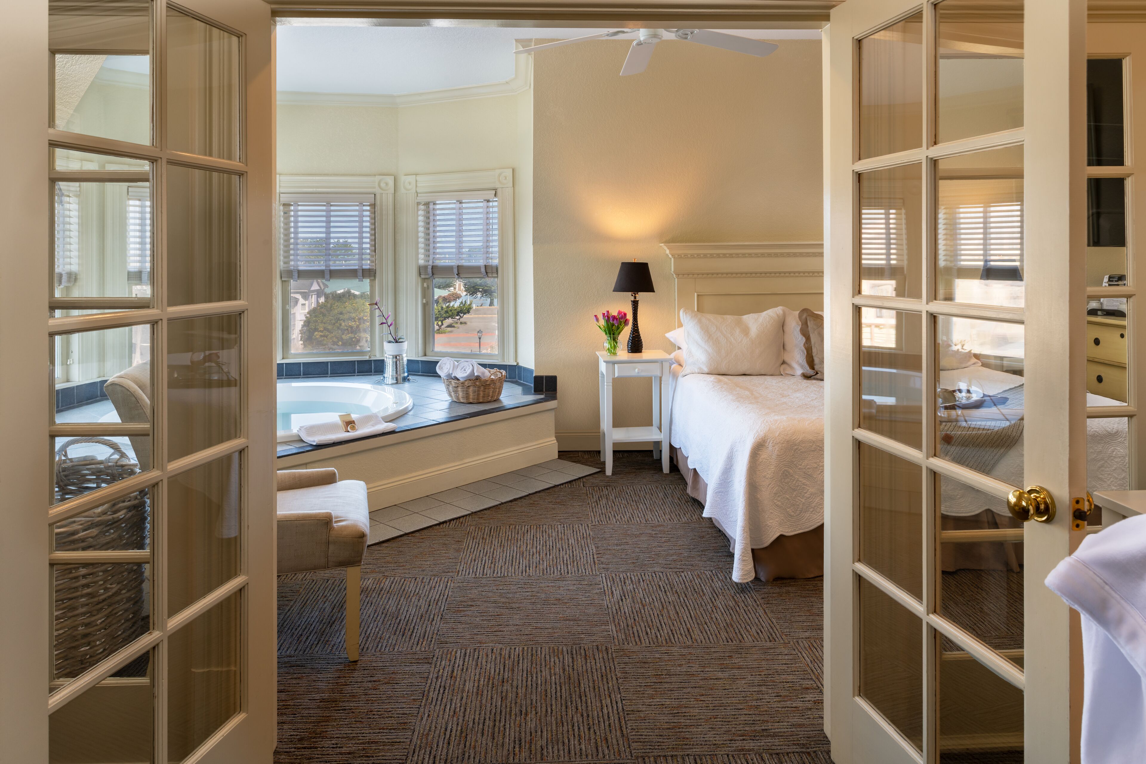 Ein Hotelzimmer mit großem Fenster, einer Badewanne, einem Bett und einem Nachttisch mit Lampe.