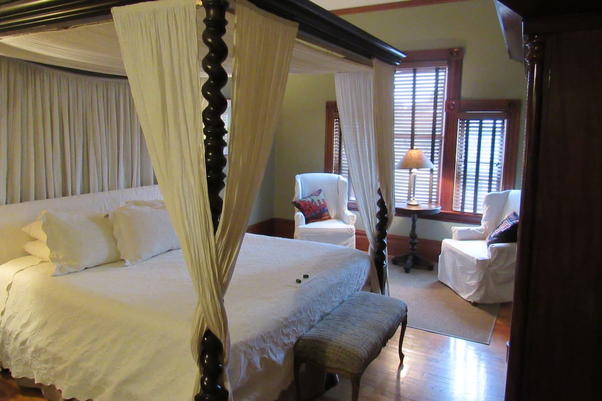 A four-poster canopy bed with a bench, a chair, and a table with a lamp in a room with wooden floors and a window with blinds.