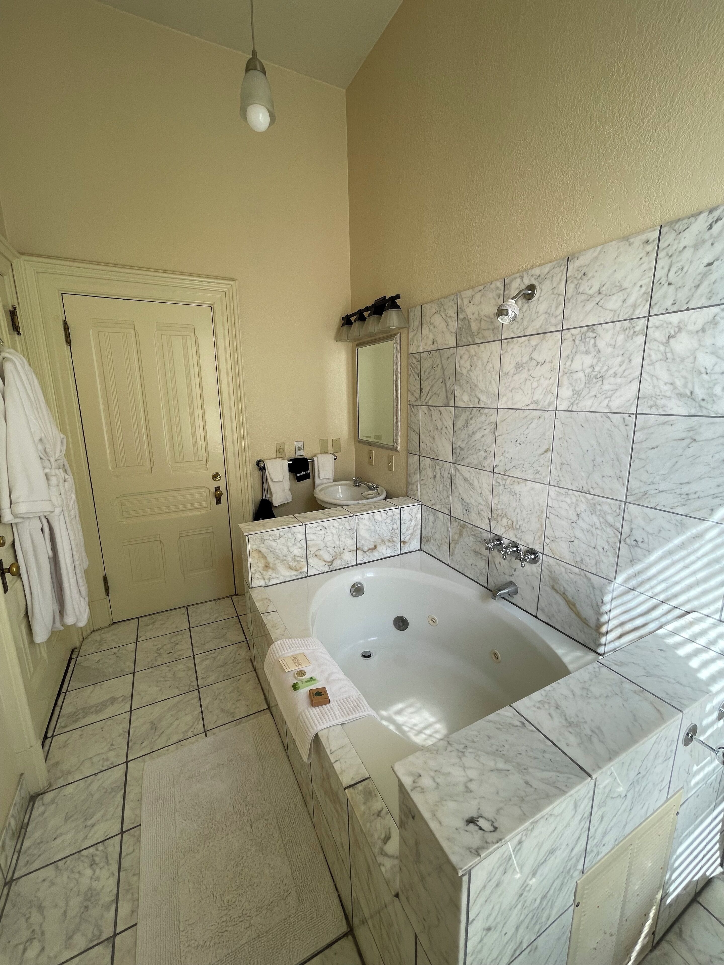 Un baño con una bañera de mármol, un lavabo y un espejo. Hay toallas colgadas en la pared y una puerta con un perchero.