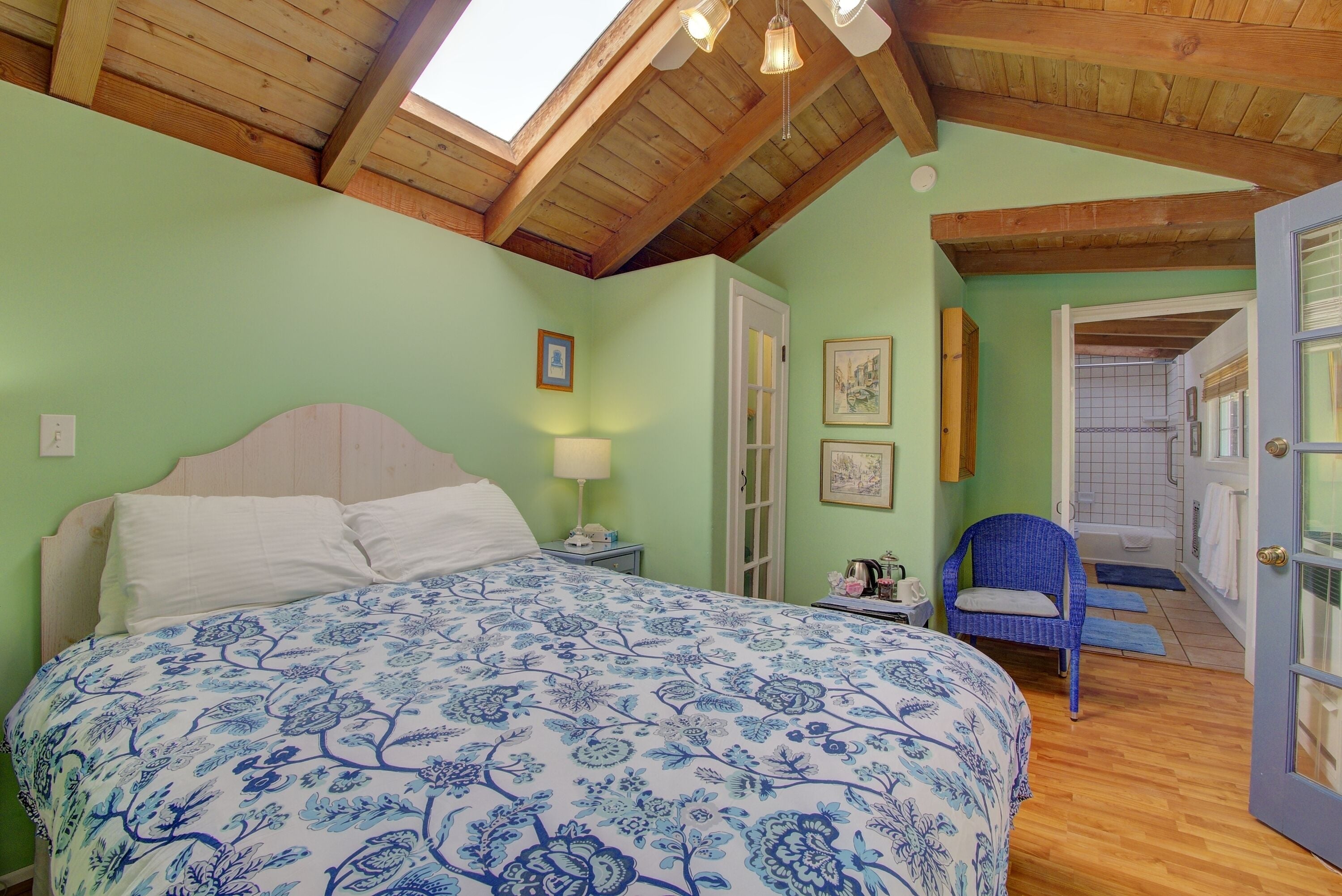 Un dormitorio con una cama grande, vigas de madera en el techo, un tragaluz, una silla y un baño visible a través de una puerta abierta.