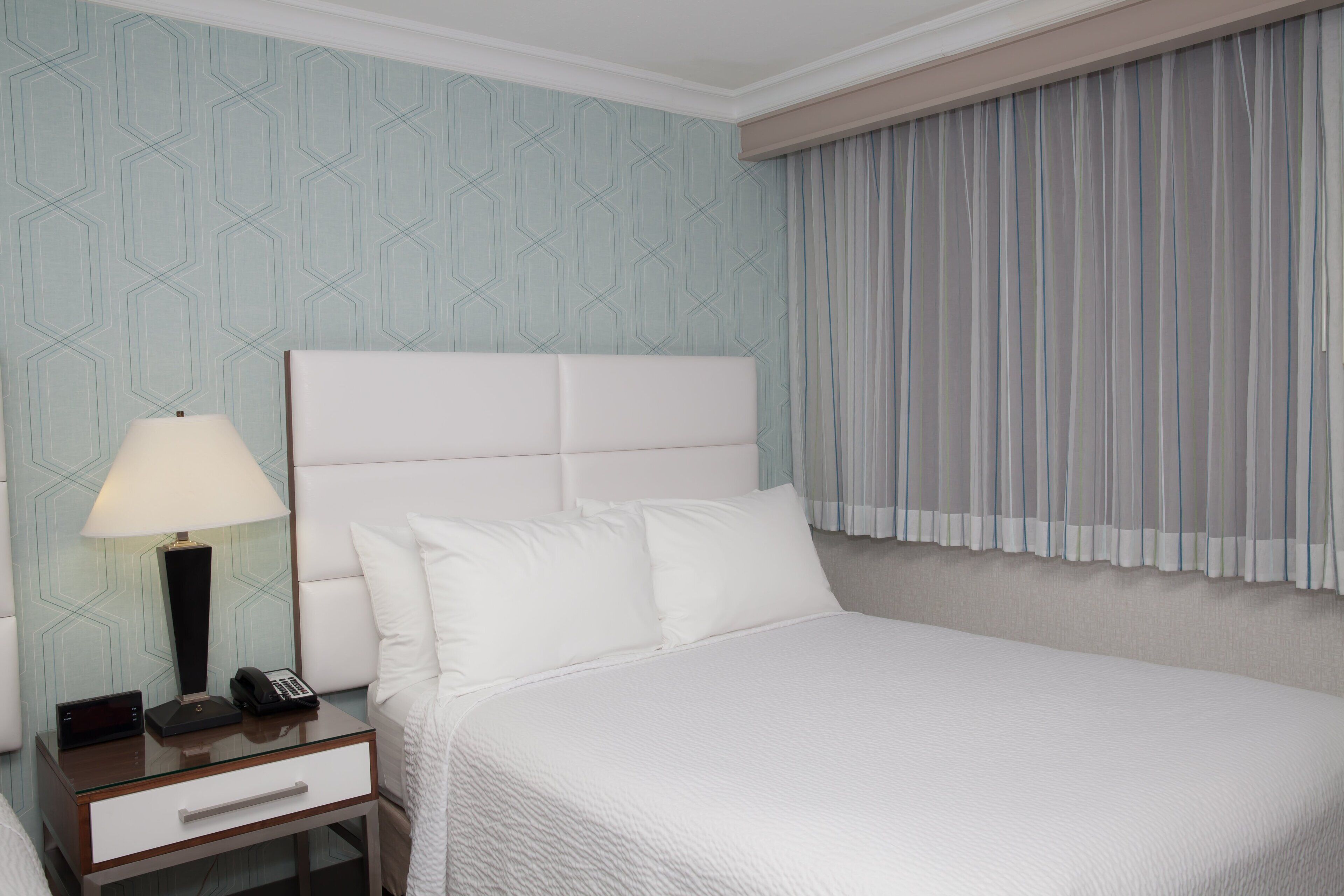 Habitación de hotel con una cama grande, una mesita de noche con lámpara, teléfono y un cajón.