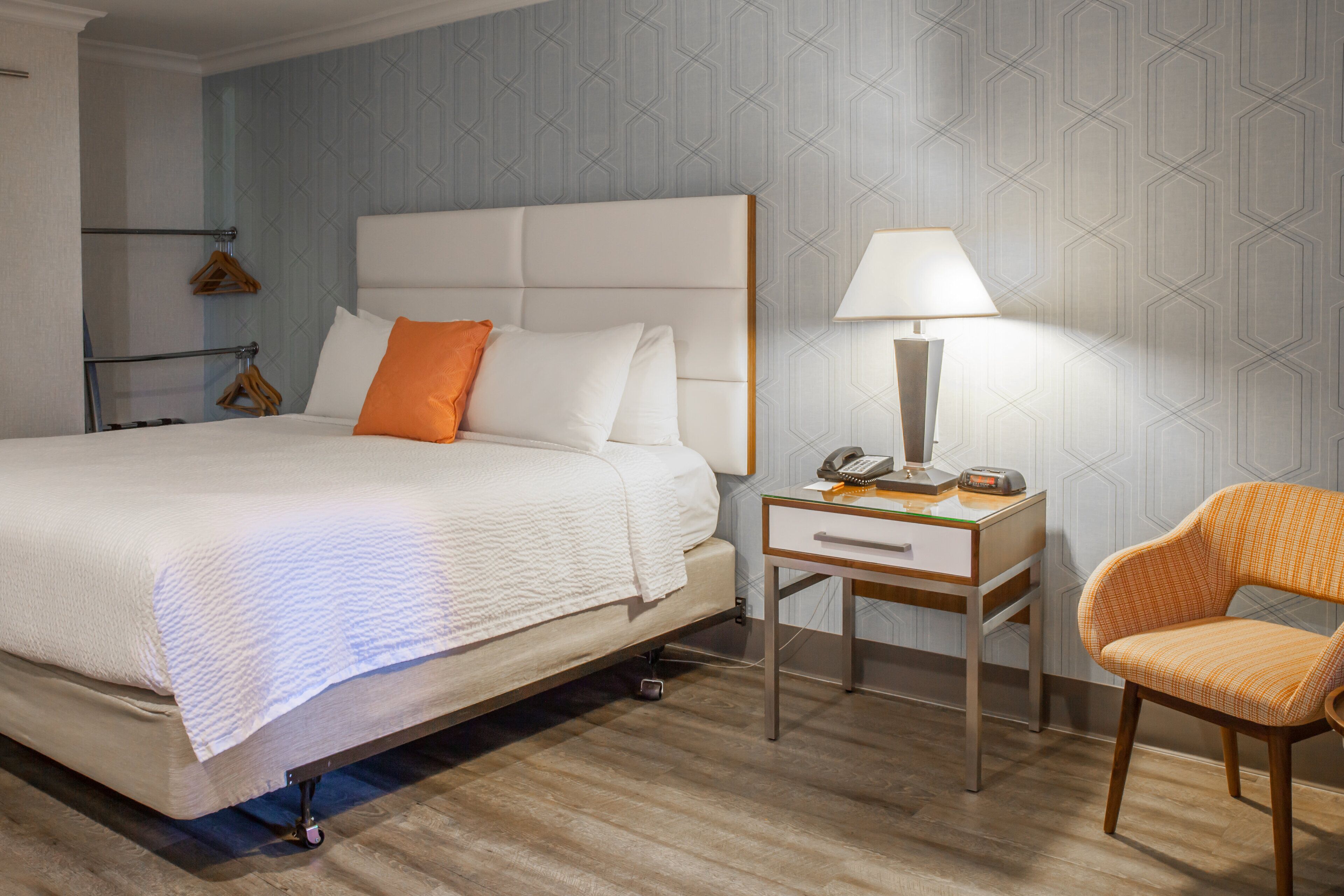 A neatly arranged hotel room with a bed, bedside table, lamp, chair, and a wall with a geometric pattern.