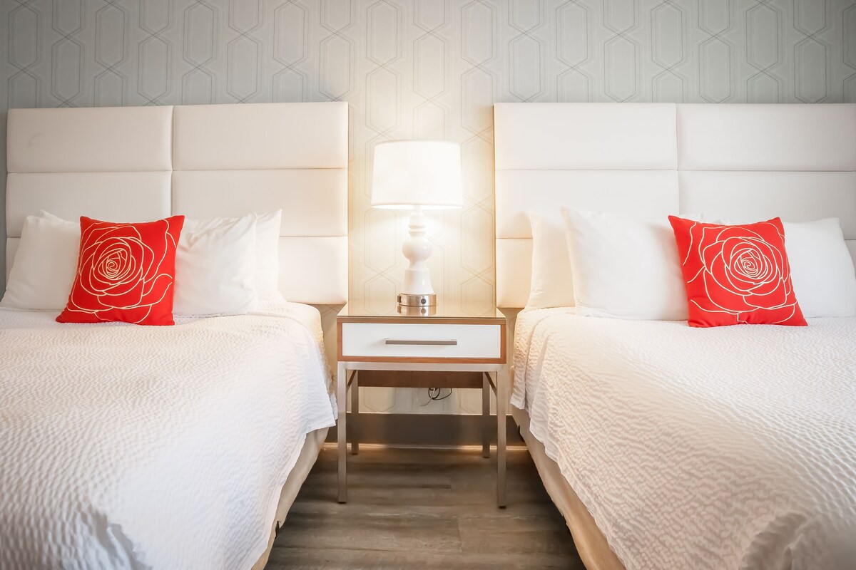 Habitación de hotel con dos camas, cada una con ropa blanca y una almohada con estampado de rosas rojas. Entre las camas hay una mesita de noche con una lámpara.