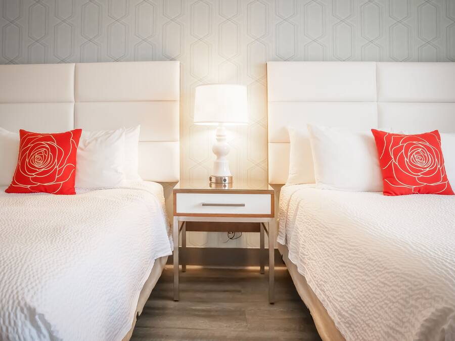 Habitación de hotel con dos camas, cada una con ropa blanca y una almohada con estampado de rosas rojas. Entre las camas hay una mesita de noche con una lámpara.