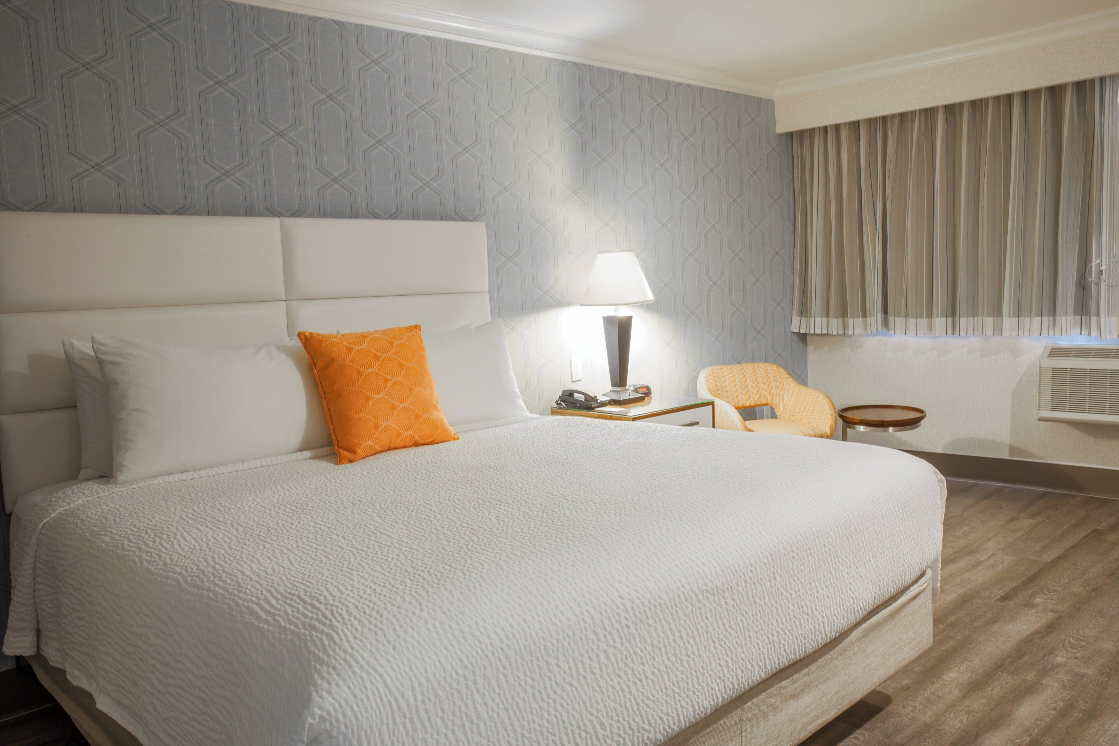 Una habitación de hotel con una cama grande, una mesita de noche con lámpara, una silla y una ventana con cortinas.
