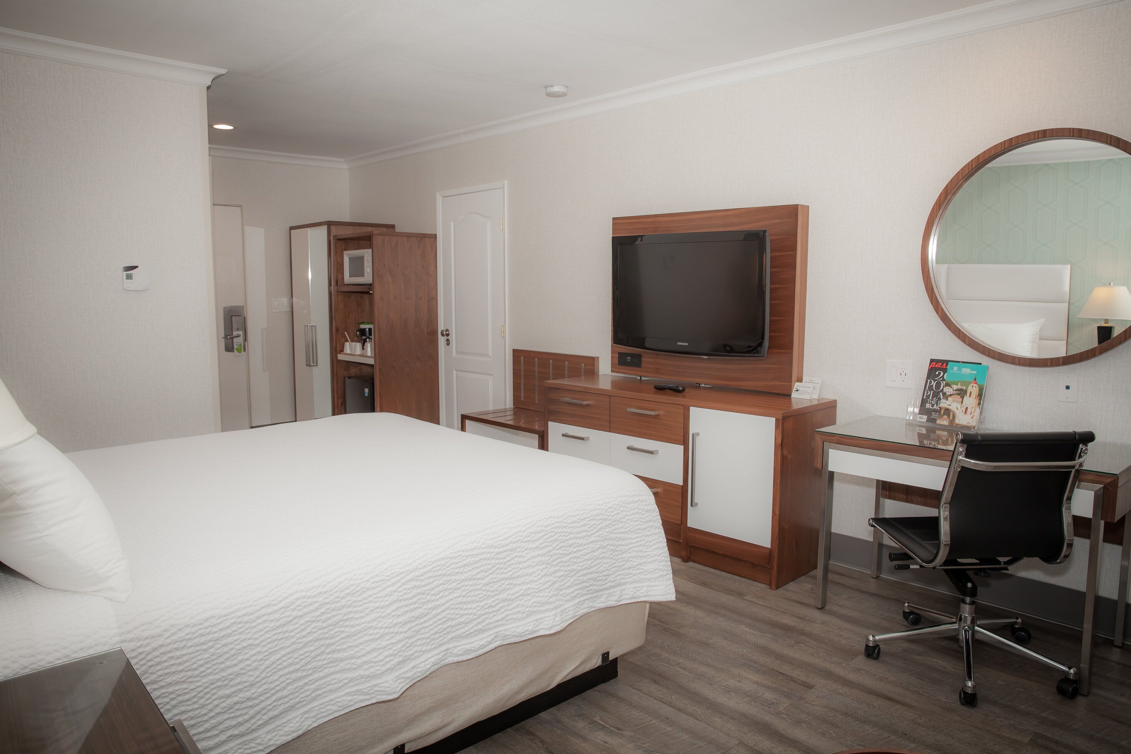 A hotel room with a bed, a desk with a chair, a television, a mirror, and a minibar.