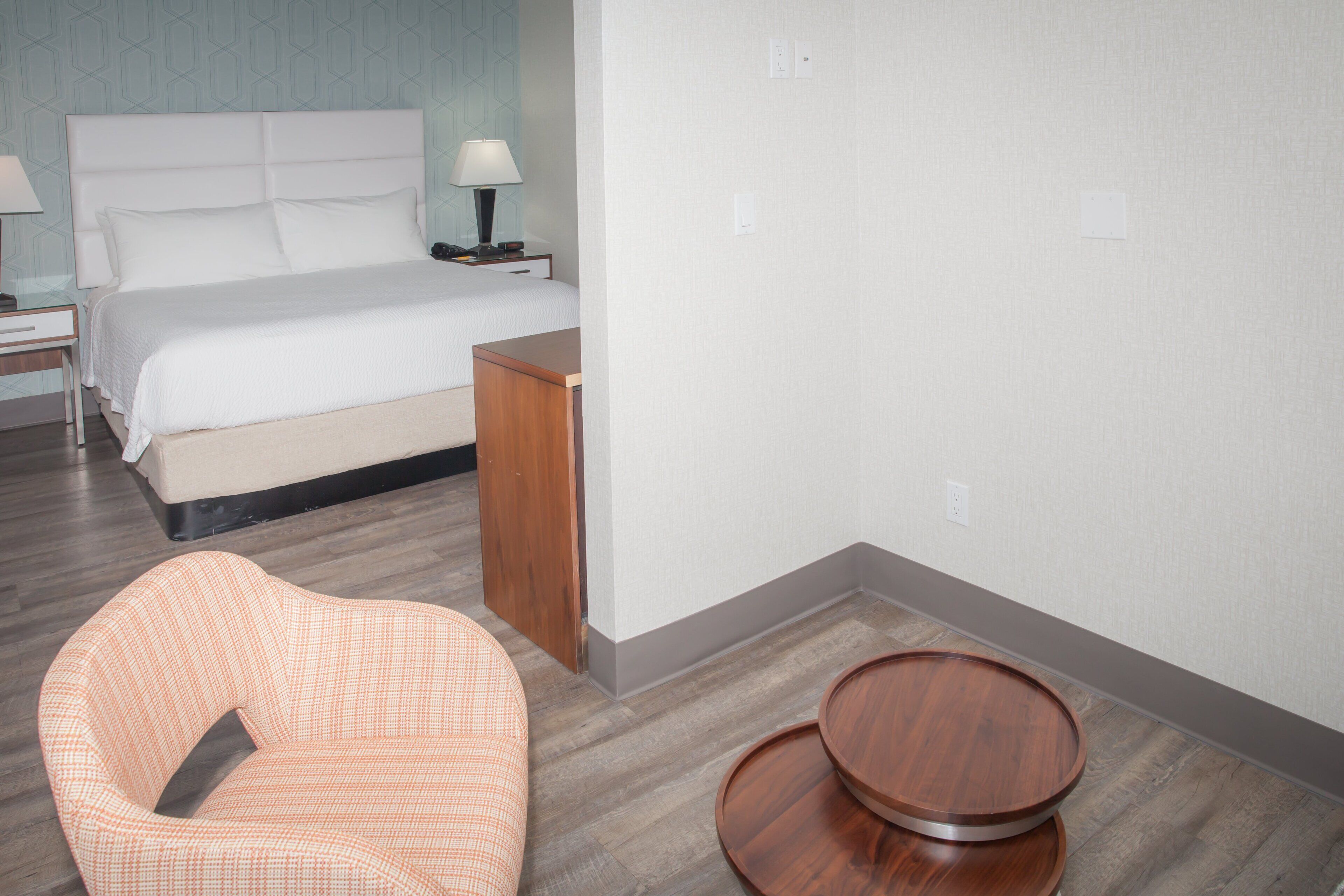 A hotel room with a bed, a nightstand with a lamp, a wooden side table, and a chair.