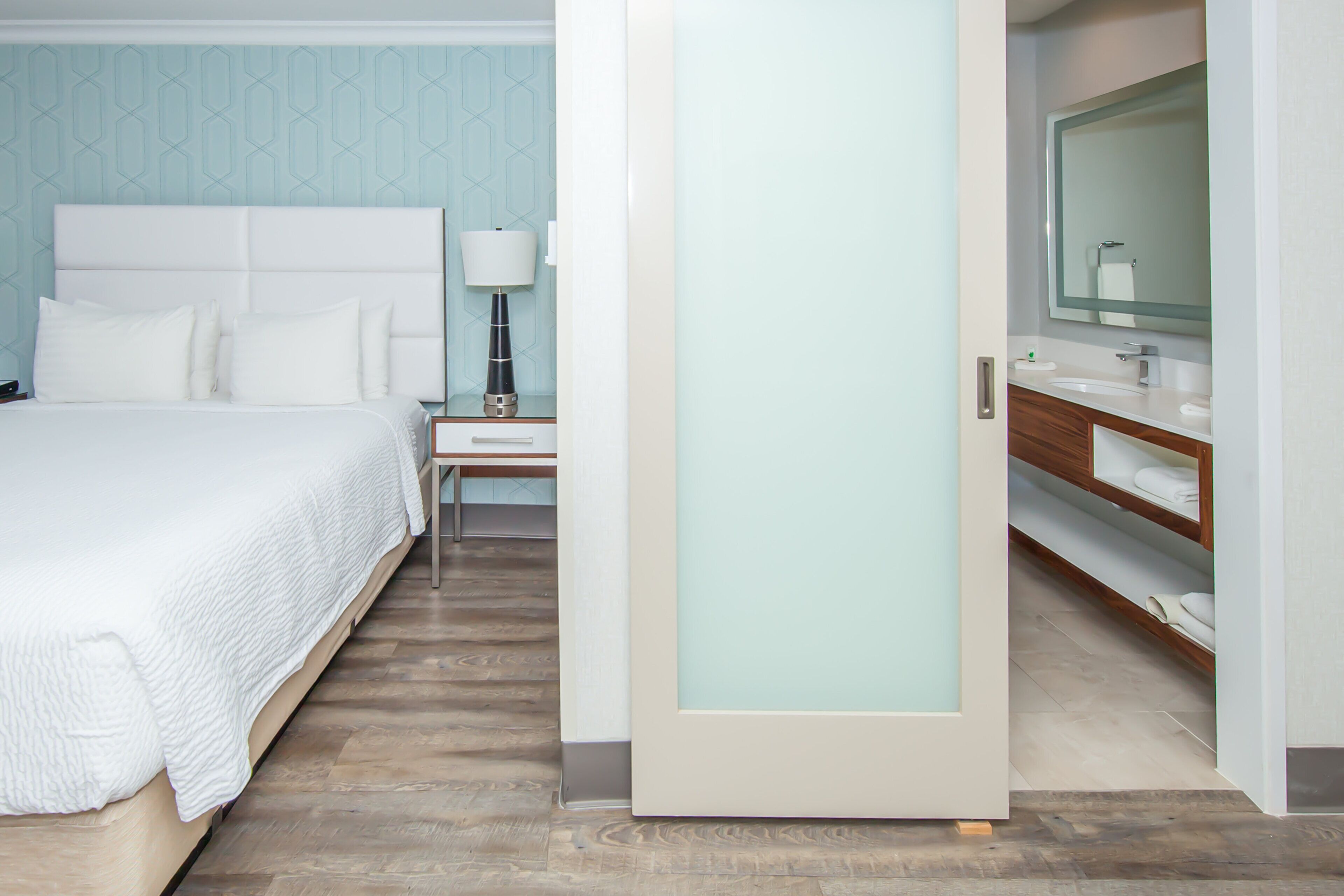 Una habitación de hotel con cama, mesita de noche, lámpara, baño con espejo y tocador de madera.