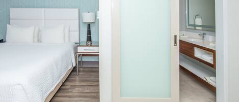 Una habitación de hotel con cama, mesita de noche, lámpara, baño con espejo y tocador de madera.