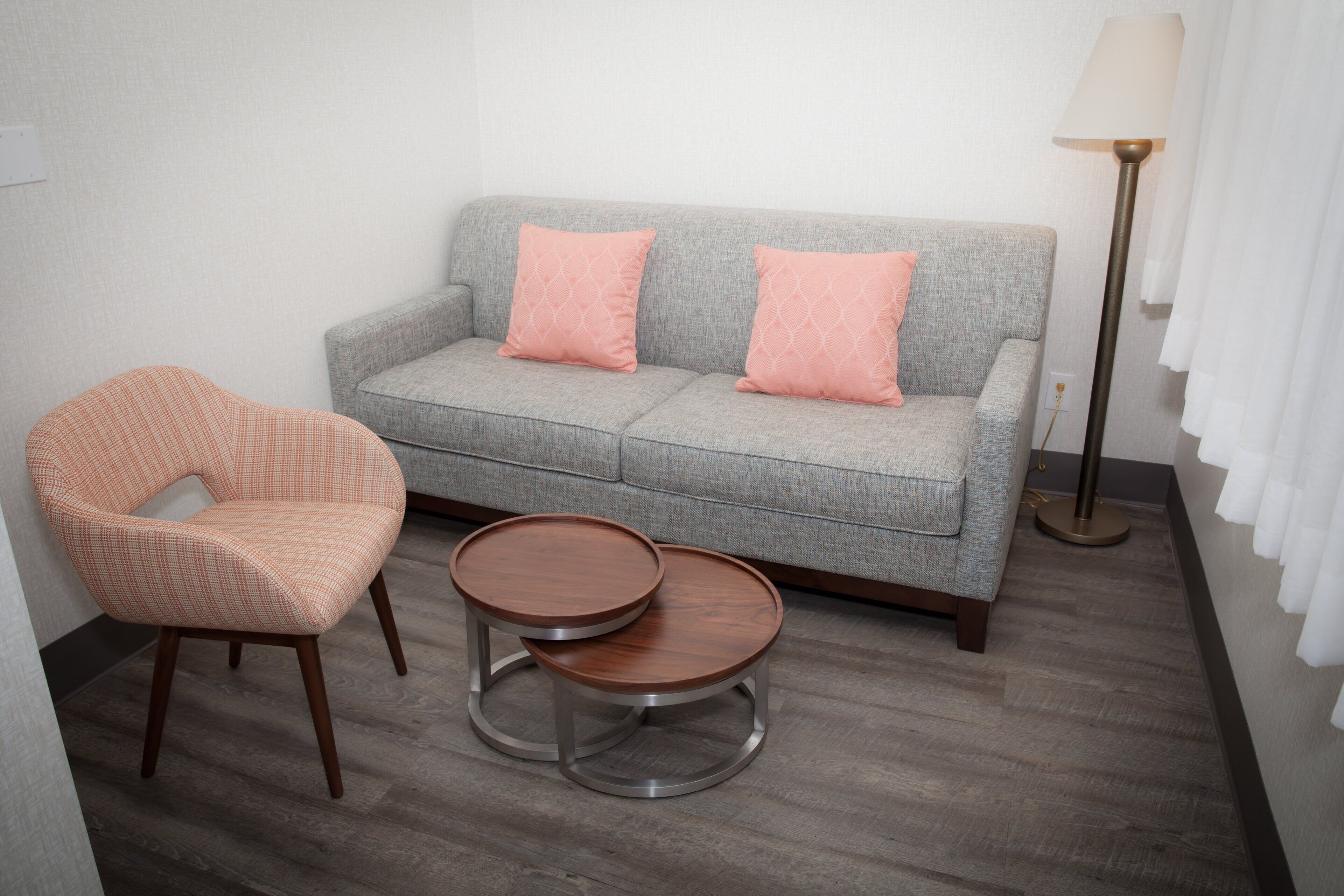 A grey sofa with two pink pillows, a wooden chair with a checkered cushion, and two round wooden tables.