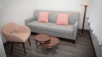 A grey sofa with two pink pillows, a wooden chair with a checkered cushion, and two round wooden tables.