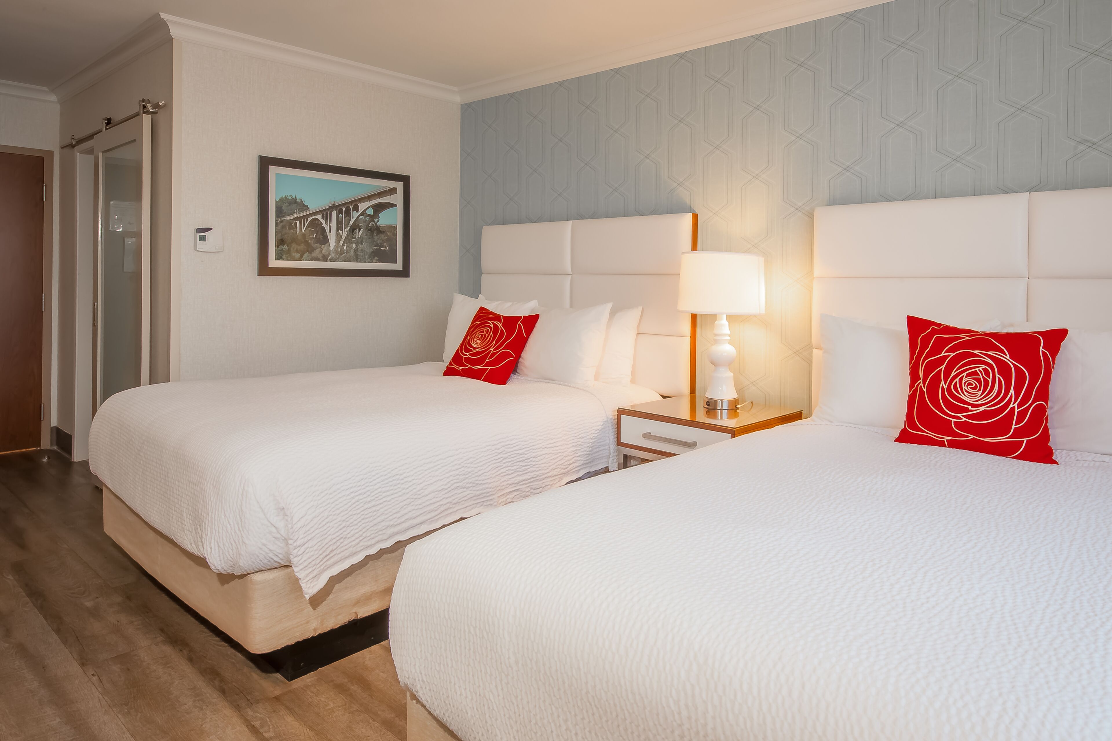 Habitación de hotel con dos camas, una mesita de noche con lámpara, un cuadro en la pared y una puerta con sistema de corredera.