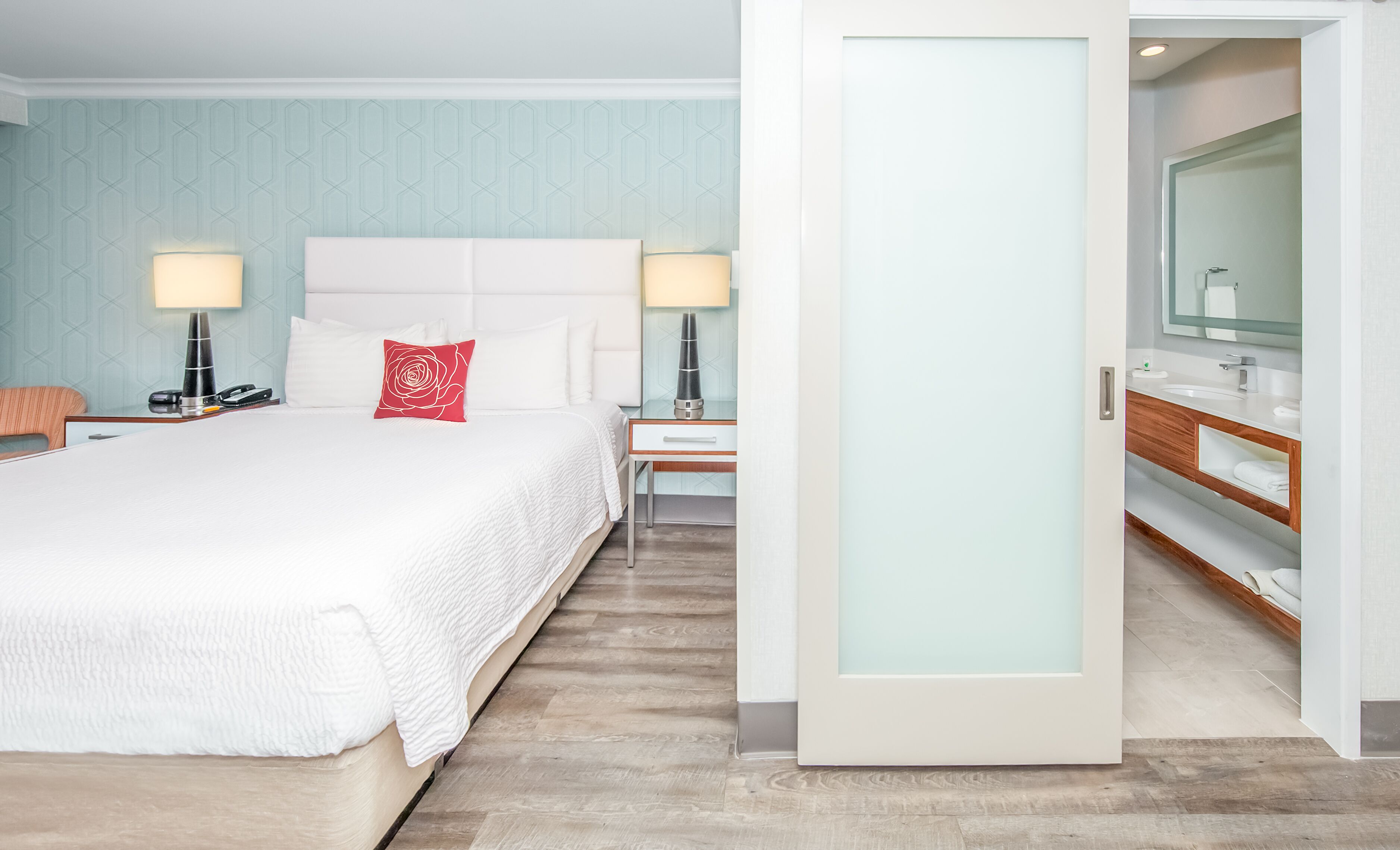 Une chambre d’hôtel avec un grand lit, des tables de chevet avec des lampes, une table de nuit, une commode en bois avec un miroir et une porte donnant accès à la salle de bain.