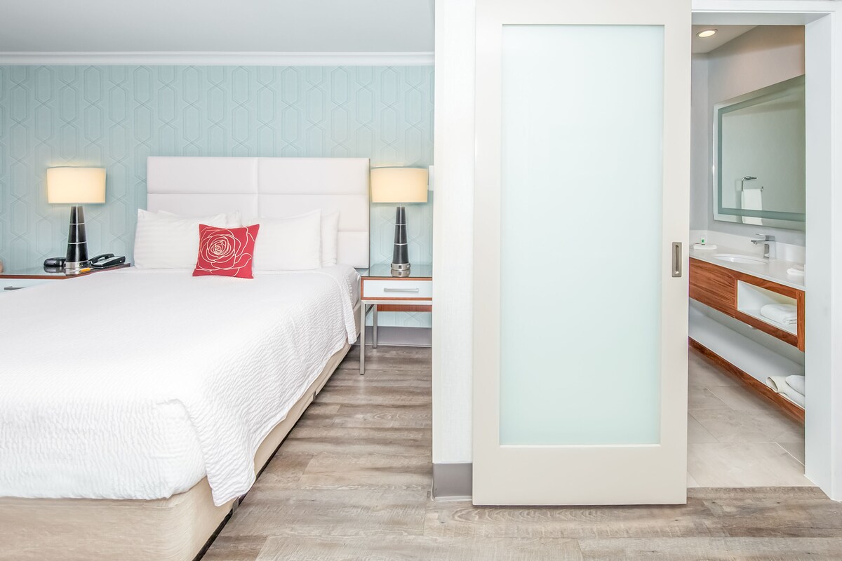 Una habitación de hotel con una cama grande, mesitas de noche con lámparas, un velador, un aparador de madera con un espejo y una puerta que da al baño.