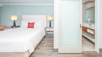 Une chambre d’hôtel avec un grand lit, des tables de chevet avec des lampes, une table de nuit, une commode en bois avec un miroir et une porte donnant accès à la salle de bain.