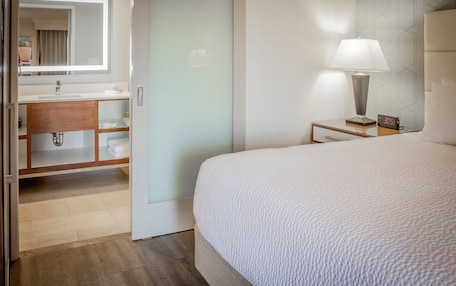 A hotel room with a bed, a bedside table with a lamp, a nightstand with a clock, a bathroom with a sink, mirror, and towel rack.