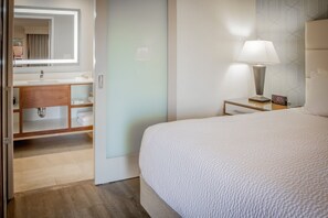A hotel room with a bed, a bedside table with a lamp, a nightstand with a clock, a bathroom with a sink, mirror, and towel rack. - Pasadena Rose & Crown Hotel (Pasadena)