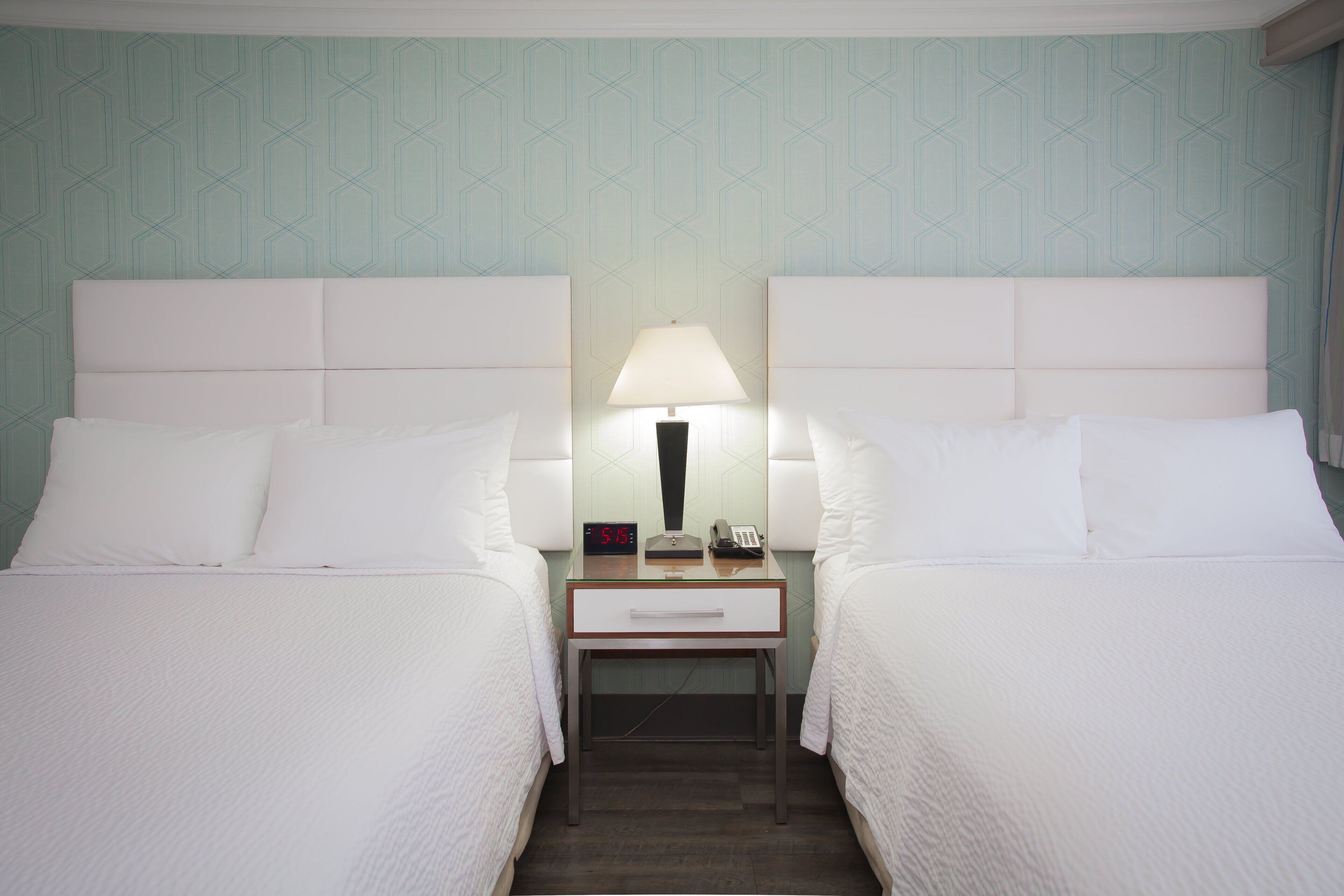 A hotel room with two beds, a nightstand with a lamp, a clock, and a phone.