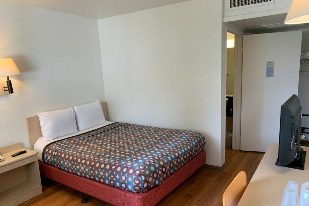A hotel room with a single bed, a bedside table with a lamp, a desk with a computer, and a chair.