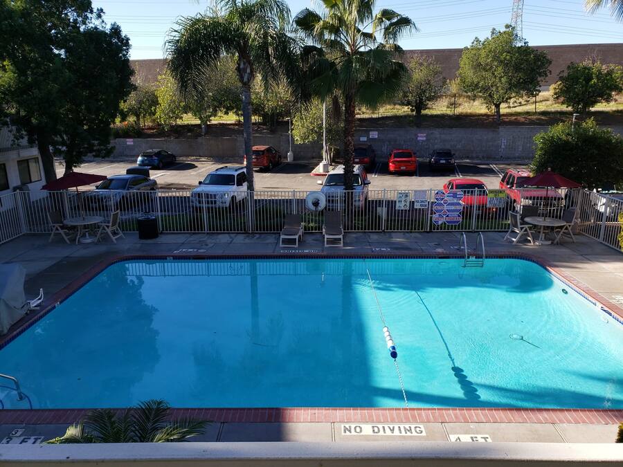 Una piscina amb un senyal de prohibició de salts, envoltada per una tanca, a prop d'una zona de pàrquing amb diversos cotxes.