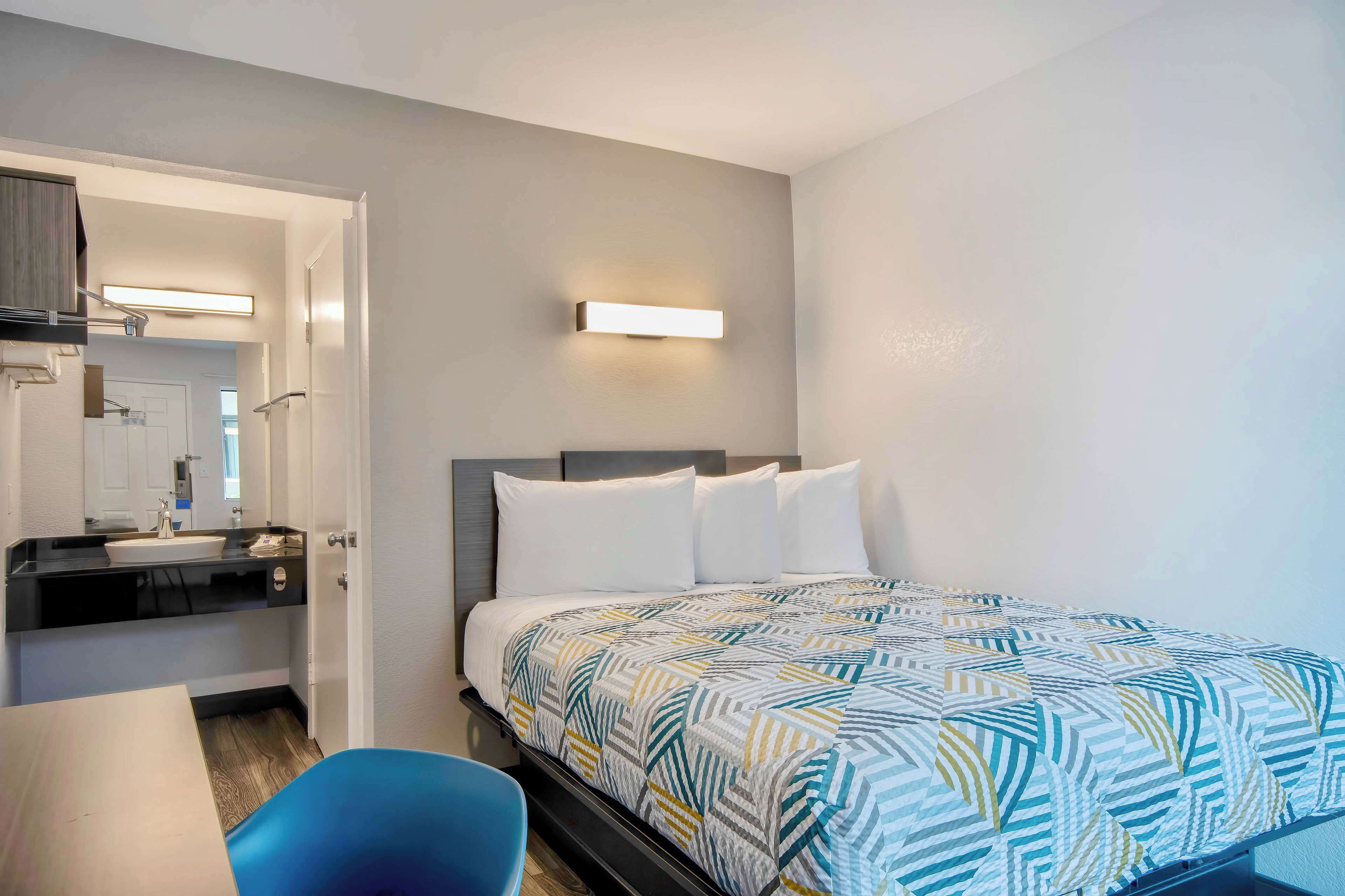 Habitación de hotel con cama, escritorio, silla, baño con espejo y una lámpara de pared.