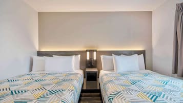 Habitación de hotel con dos camas, una mesita de noche con lámpara y una pared lisa.