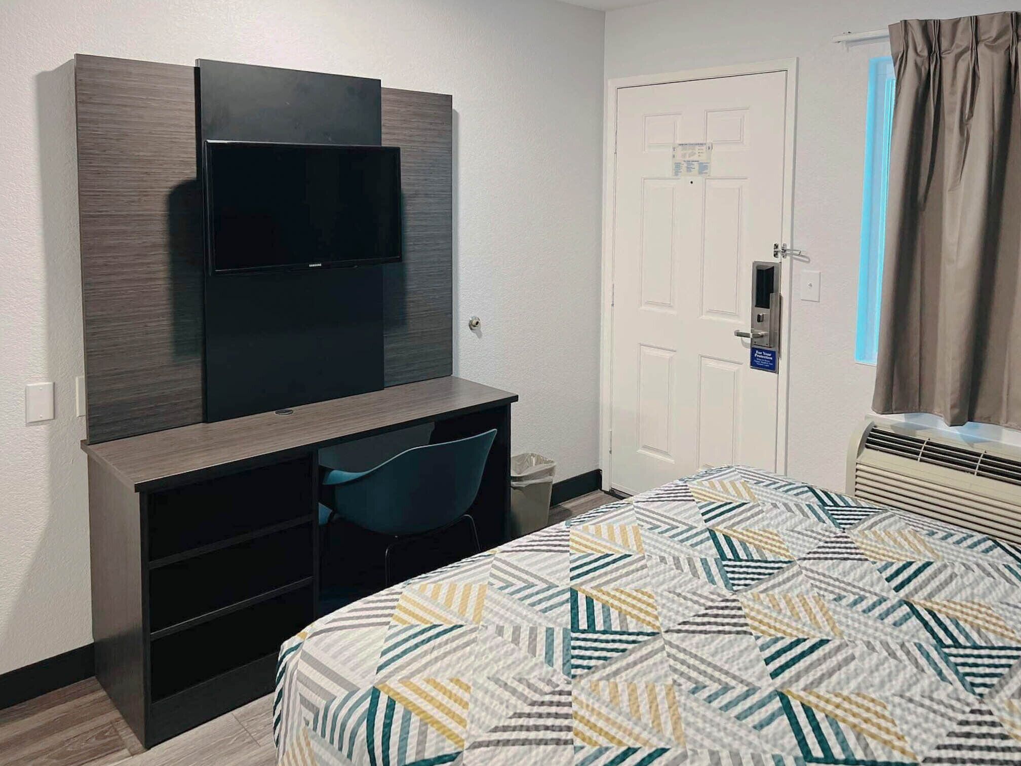 A hotel room with a bed, a desk with a chair, a television mounted on the wall, a door with a sign, and an air conditioning unit.
