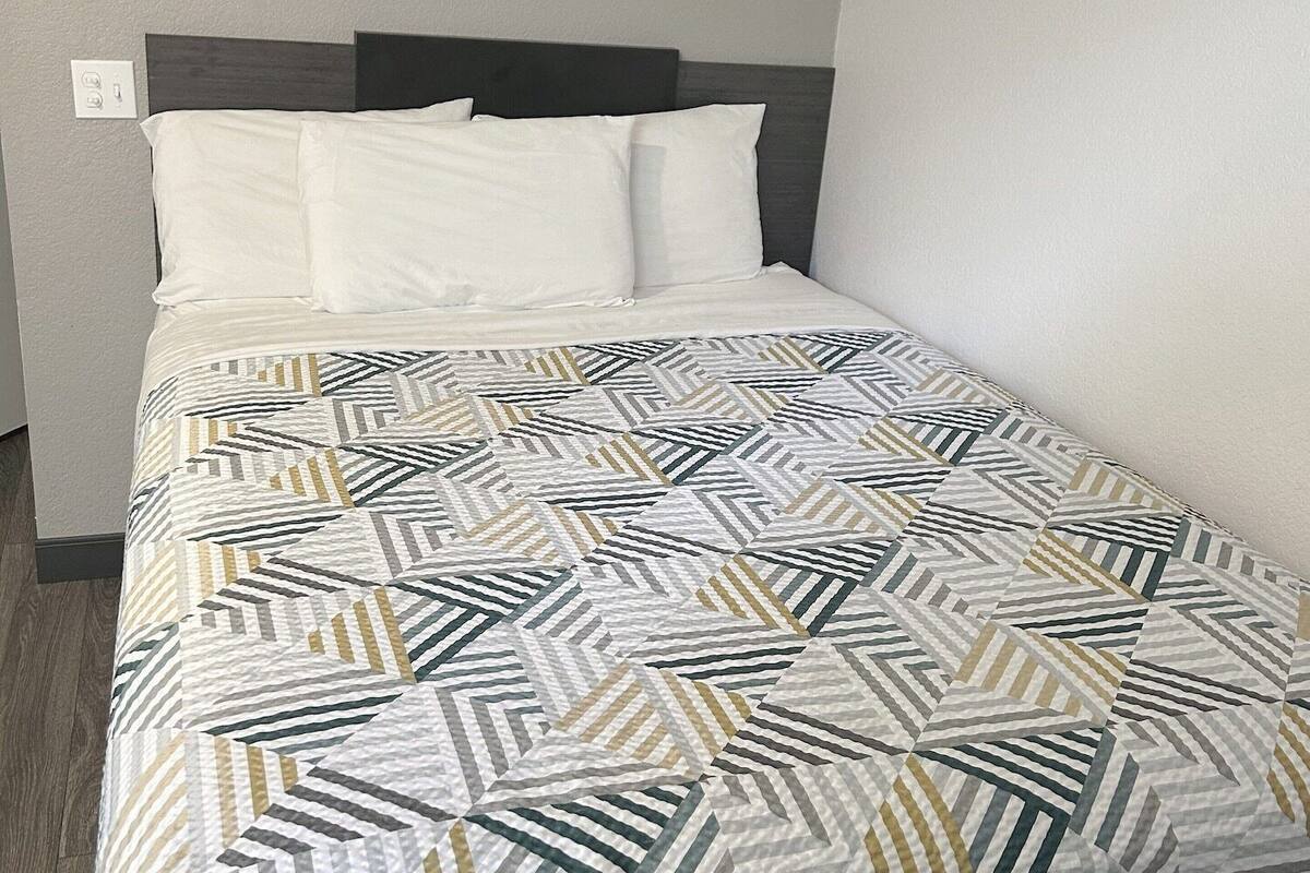 Ein ordentlich bezogenes Bett mit einem geometrisch gemusterten Überwurf, einem dunklen Kopfteil und einer schlichten Wandleuchte.