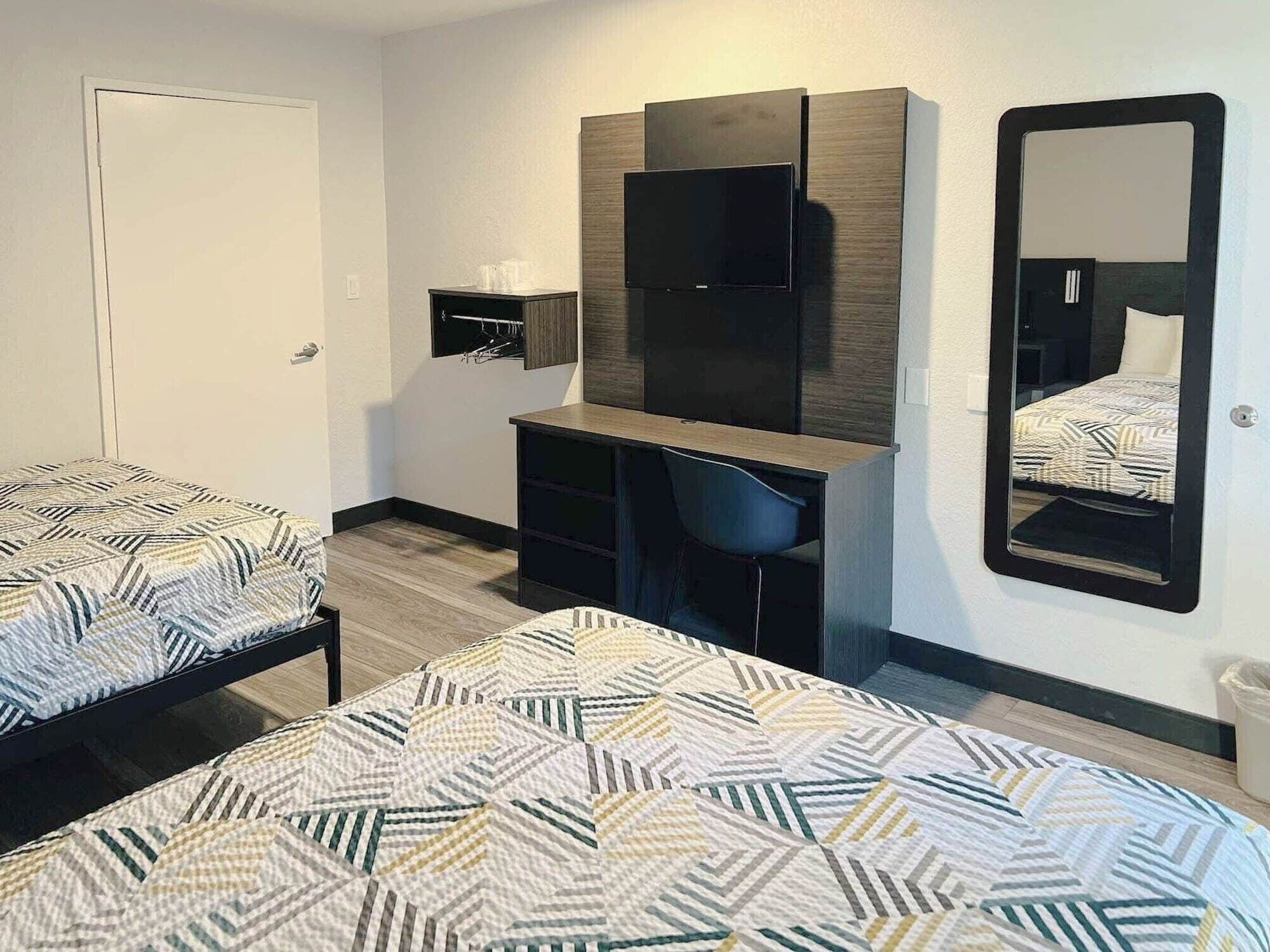 Quarto de hotel com duas camas, uma escrivaninha, uma cadeira, um guarda-roupa, um espelho e uma pequena prateleira com artigos de higiene pessoal.
