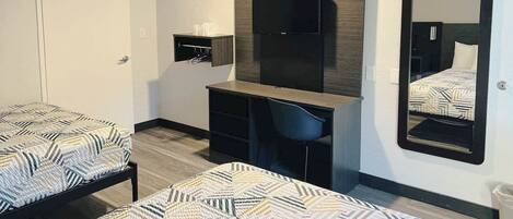 A hotel room with two beds, a desk, a chair, a mirror, and a small shelf with toiletries.