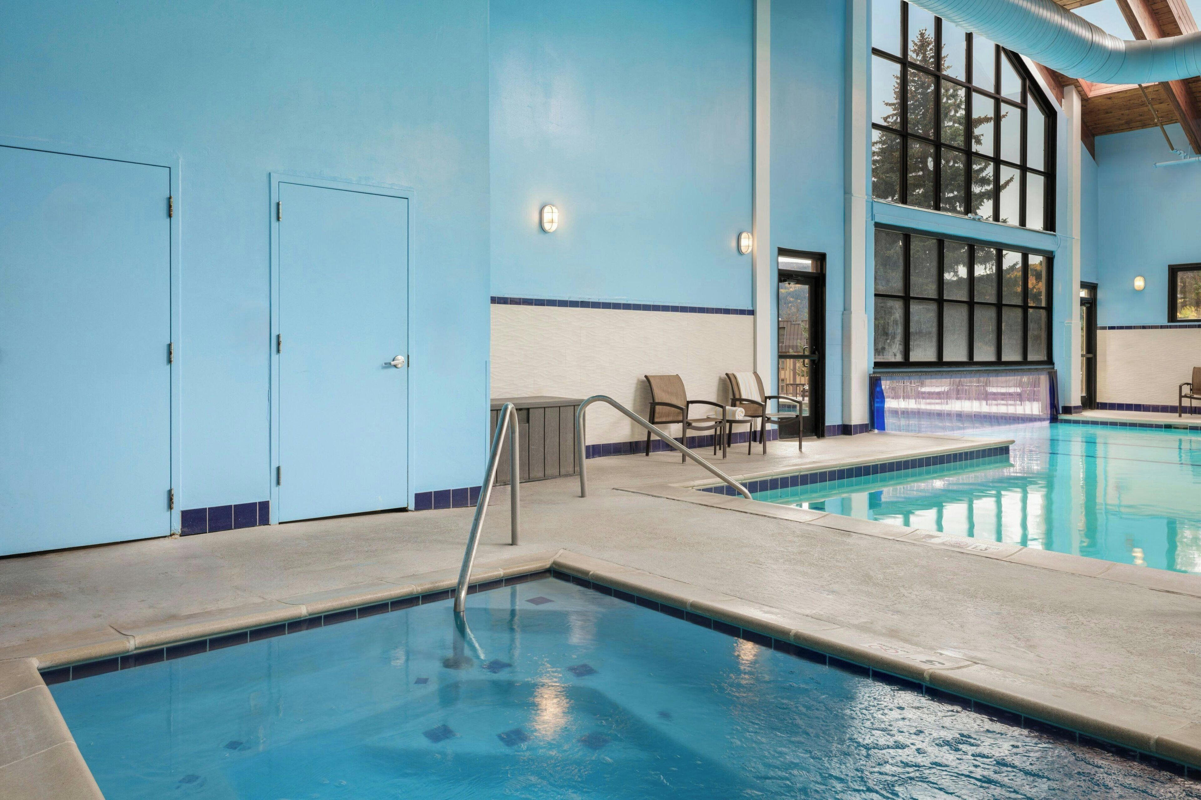 An indoor swimming pool with clear blue water, a tiled floor, and a seating area with chairs.