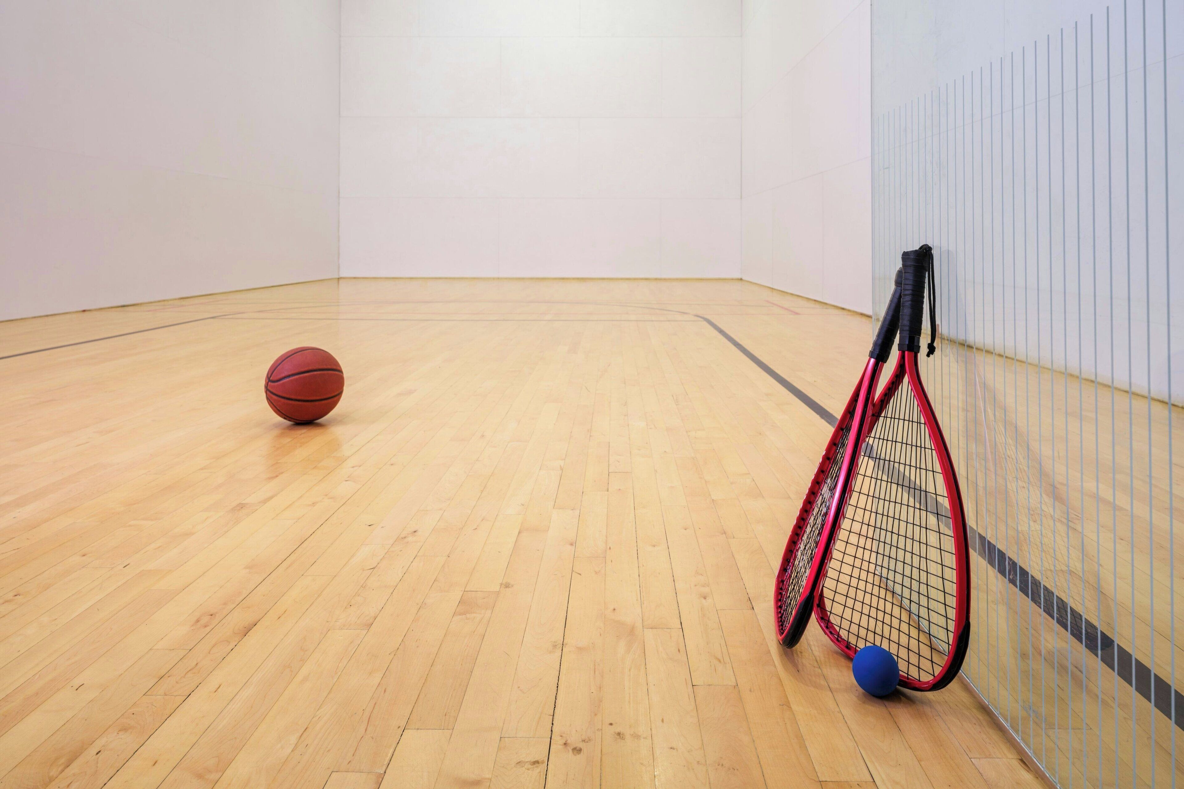 An indoor sports court with a basketball and a tennis racket leaning against the wall.