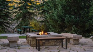 A stone fire pit with benches, surrounded by a stone patio and a view of trees and a mountain in the background. - Marriott's StreamSide Evergreen at Vail (Vail)