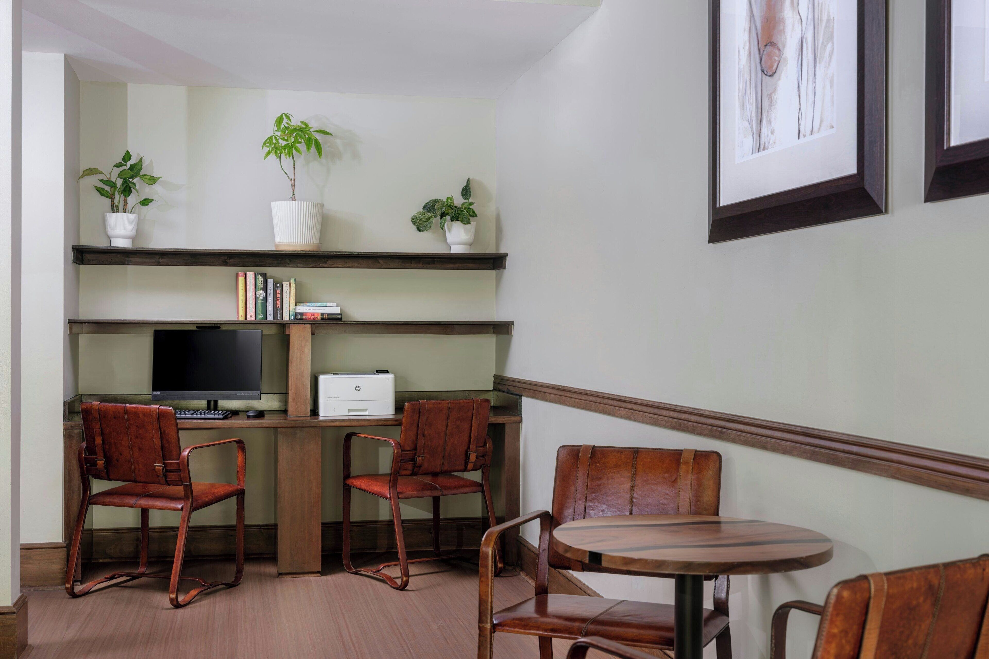 A modern office space with a desk, two chairs, a round table, and three potted plants on shelves.