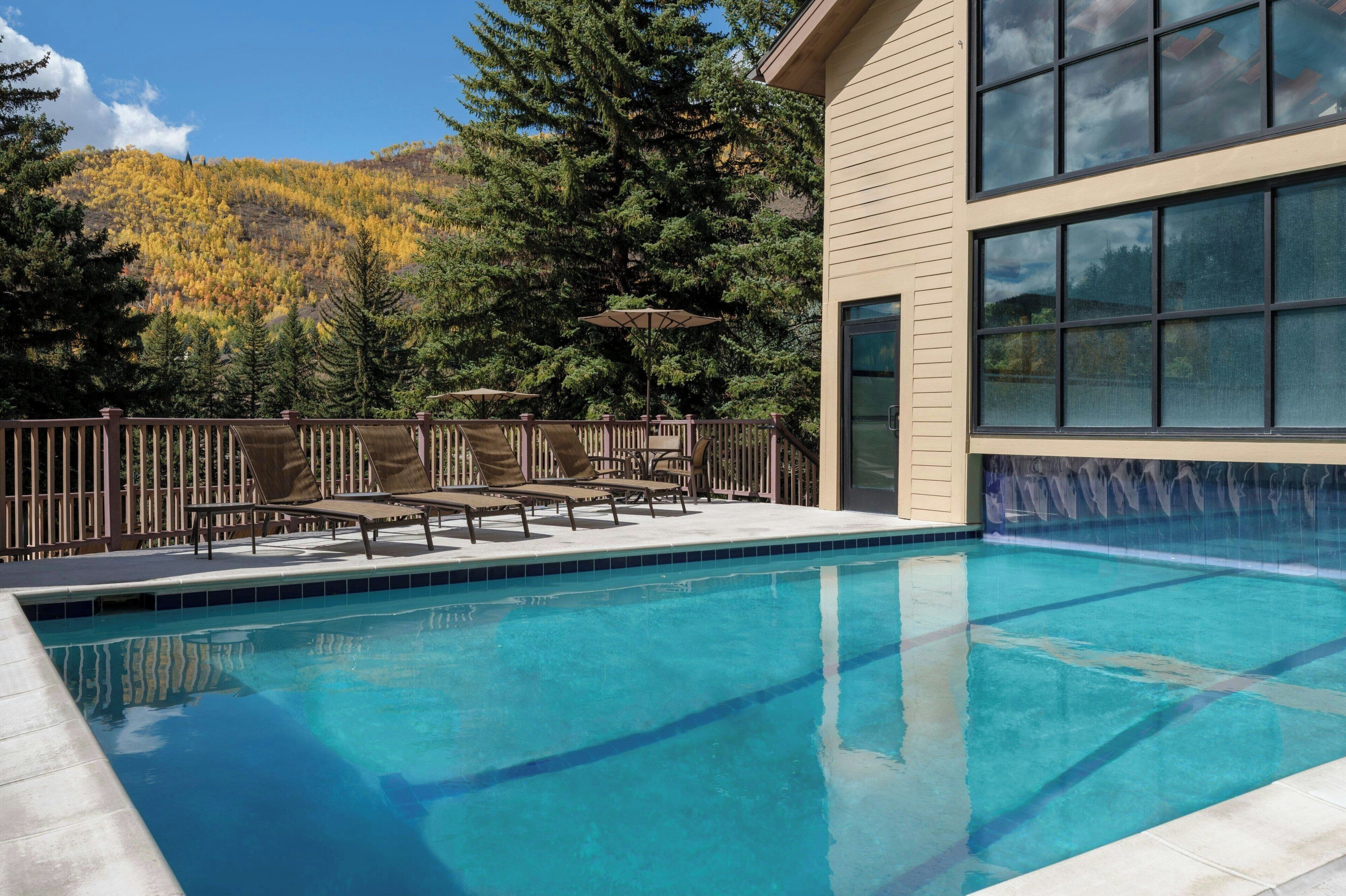 Piscina con sillas de descanso y sombrillas, rodeada por una terraza de madera y un paisaje montañoso.